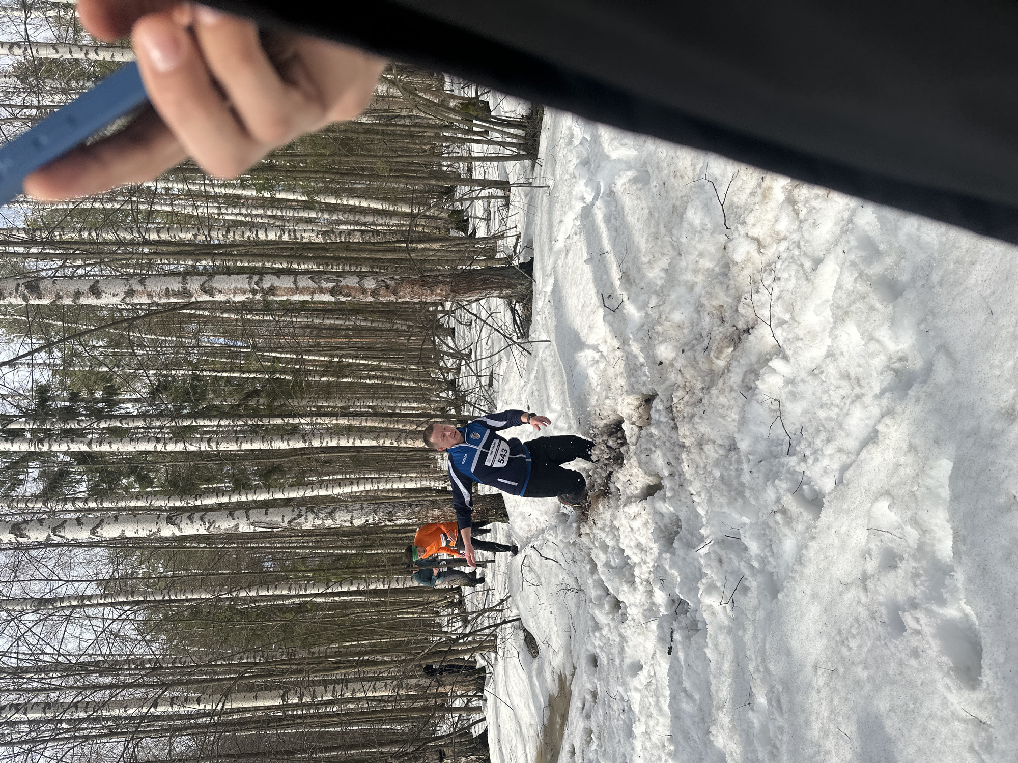 The image depicts a snow-covered landscape with birch trees in the background. A man wearing a blue and black jacket with the number "543" is partially submerged in a patch of muddy snow. Other people wearing sports clothes and headbands appear to be present in the background, near the trees. A hand holding a blue object and a black object are also visible in the foreground. The location is Zelenodol'sk, Russia, based on other information. The setting suggests a winter sporting event or activity.