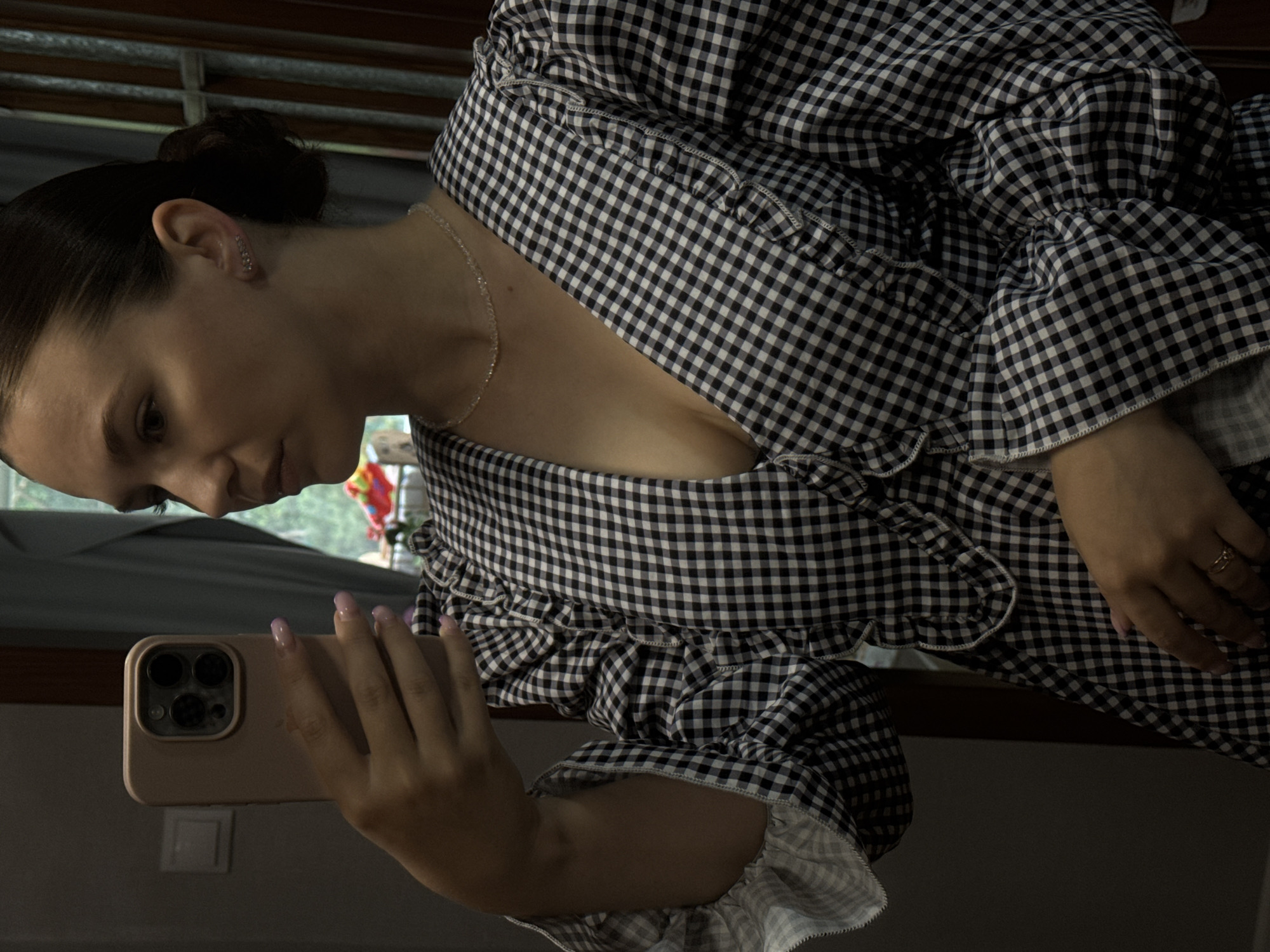 The image shows a young woman with her hair pulled back in a bun, taking a mirror selfie with her phone. She is wearing a black and white gingham dress with ruffled details on the sleeves and neckline. She has on a delicate silver necklace and stud earrings. Her nails are painted a light purple. The background appears to be an indoor setting, possibly a bedroom, with a window visible on the right side of the frame. The scene does not provide any specific indicators of the city of Chaikovsky, Russia.