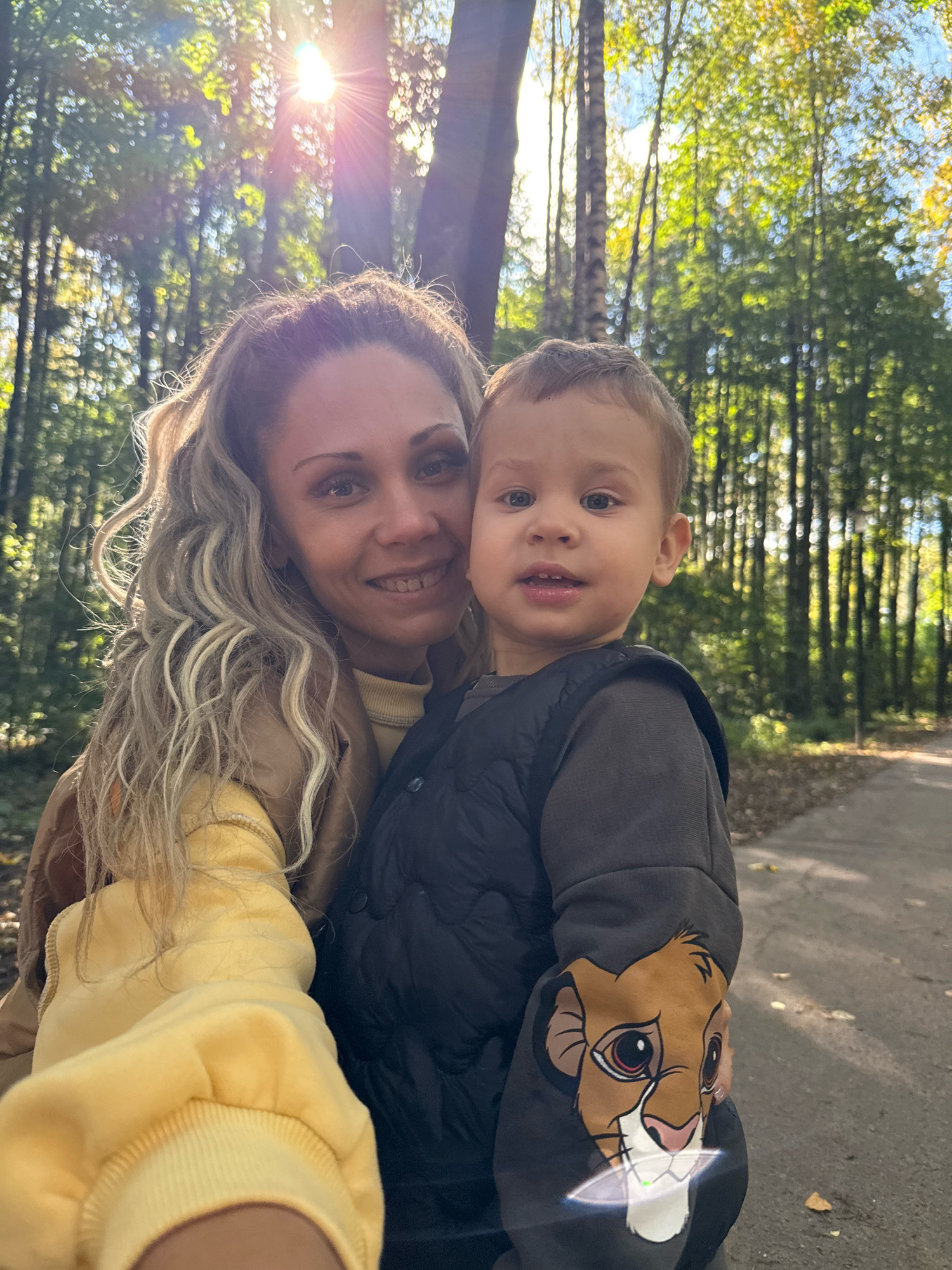 A woman with long, blonde, wavy hair and a yellow hoodie is taking a selfie with a young boy in a grey vest and a grey sweatshirt with a lion graphic on the sleeve. They are in a park in Rusanovo, Russia, with trees and sunlight in the background. The path in the park is visible in the background as well. The woman is smiling at the camera, and the boy is looking at the camera with a neutral expression.