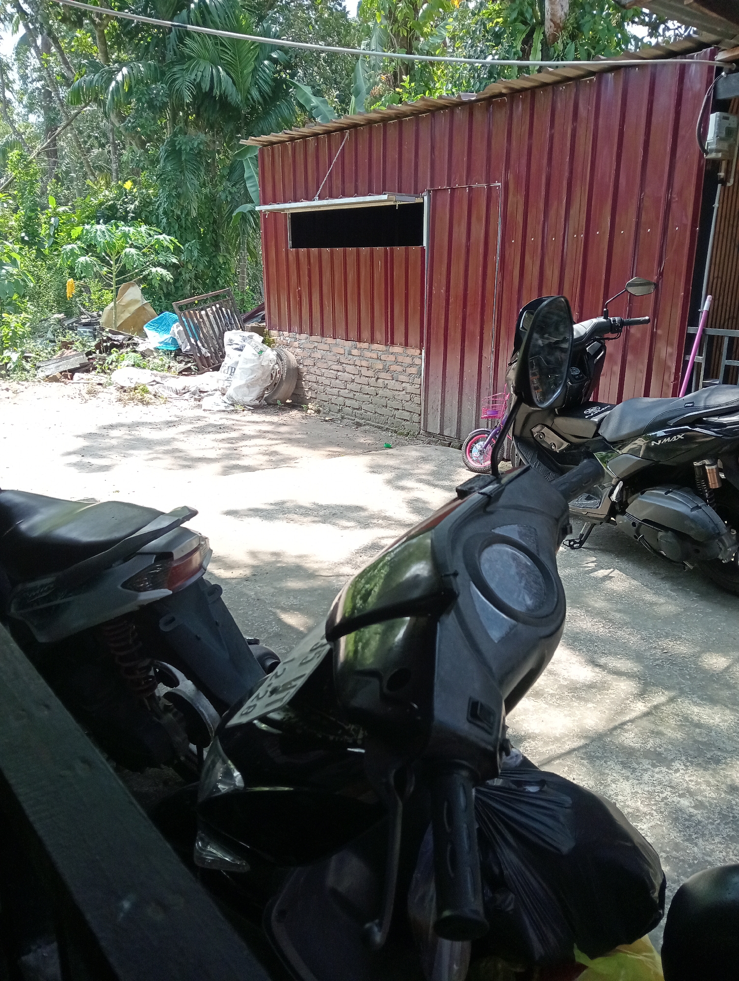In Campago, Indonesia, an outdoor scene captures a street with two parked motorcycles. The first motorcycle, black with silver accents, is in the foreground, partially obscured by what appears to be a dark tarp or bag. Its handlebars, mirrors, and front portion are visible. Behind it, a second black motorcycle, identified by "NMAX" branding on its side, is also parked. This motorcycle has a side stand engaged and its rear tire and exhaust system are visible. A small pink bicycle with a basket is positioned in front of the second motorcycle. The background features a red corrugated metal structure with a dark rectangular opening, possibly a window or vent, and a brick lower section. To the left of the structure, a pile of debris including white sacks, a blue tarp, and wooden planks is scattered amongst lush green foliage, indicating an overgrown or untidy area. Sunlight casts dappled shadows on the concrete ground. A power meter is mounted on the right side of the red structure.