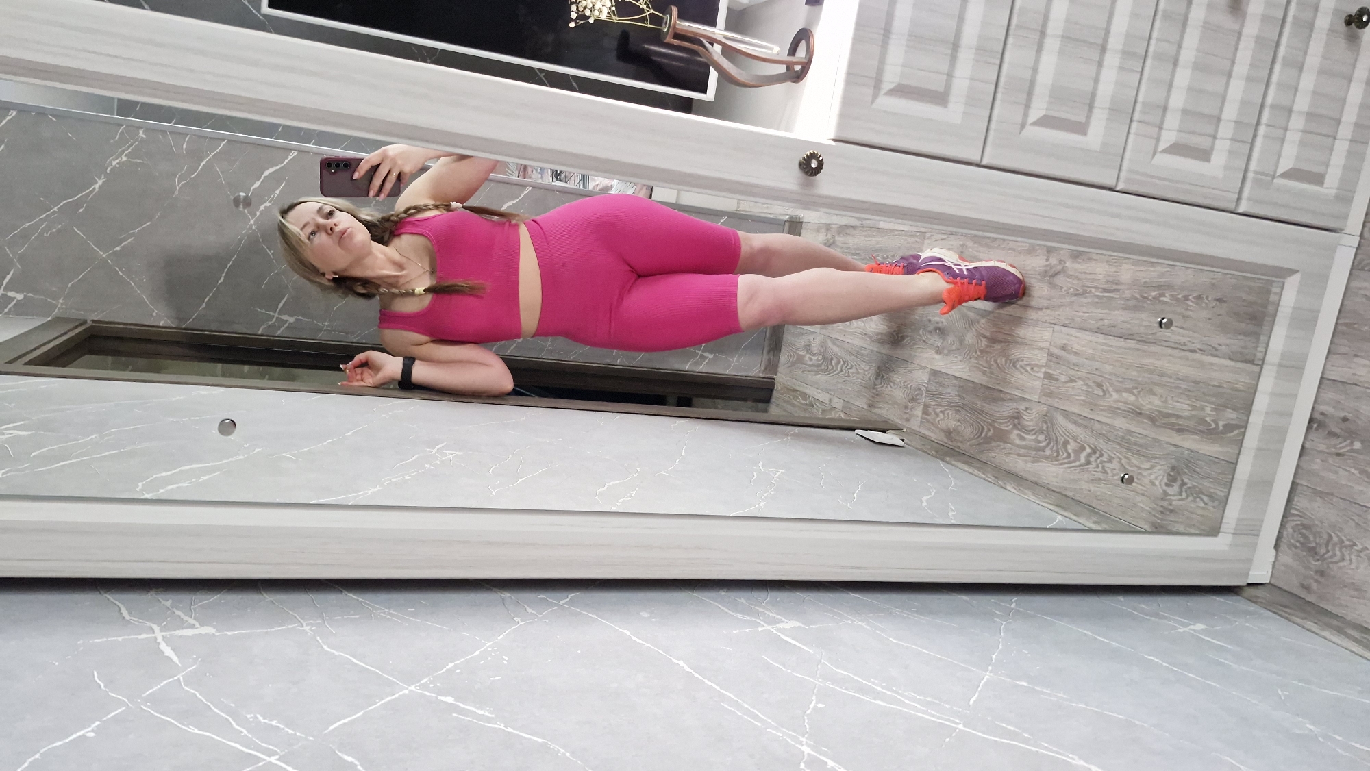 A woman is reflected in a long mirror. She is wearing a pink workout set and orange and purple athletic shoes. The floor is a light gray marble pattern, while the floor behind her is a light brown wood grain. There are cabinets above and to the right of her. The location is likely a bathroom, though the city and country are not discernible from the image.