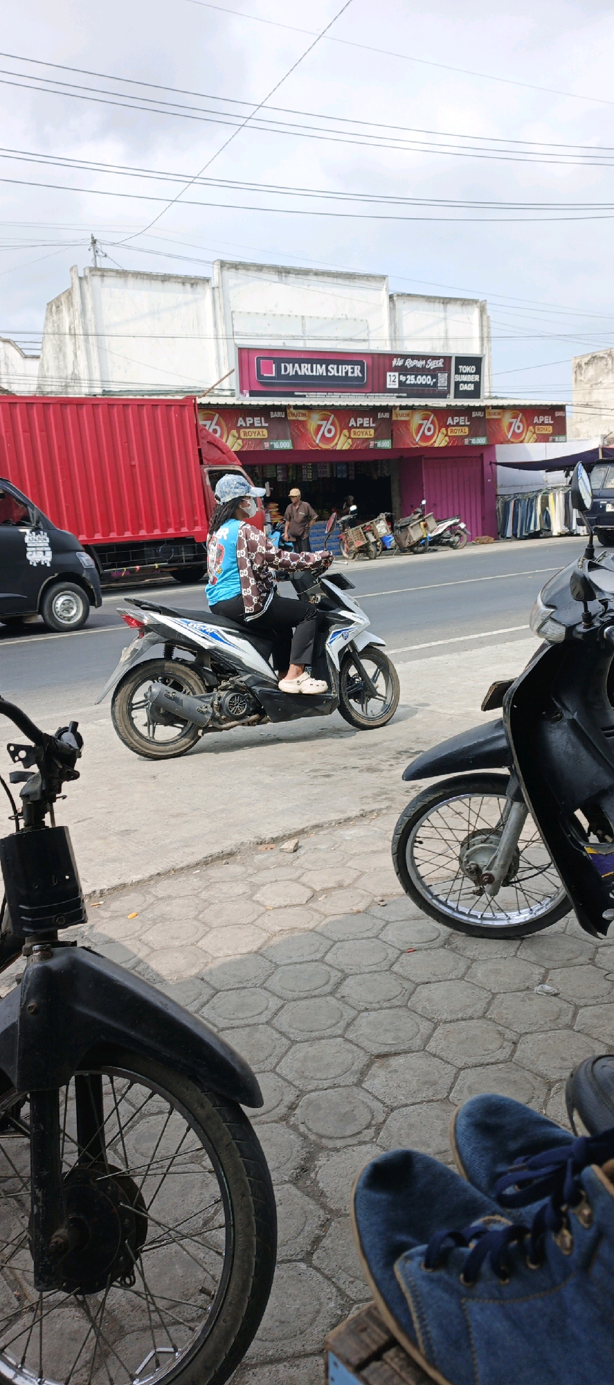 A person wearing a patterned jacket and a light blue t-shirt is riding a white and blue motorcycle on a street in Kasiyan, Indonesia. The person is also wearing a white helmet and a light blue cap. In the background, there is a "Djarum Super" store with several advertisements for "76 Apel Royal." A red truck and a black van are parked on the side of the road. Several other motorcycles are parked in front of the store. The ground is paved with hexagonal tiles, and there are power lines overhead. Two pairs of blue shoes are visible in the foreground, suggesting someone is sitting nearby.