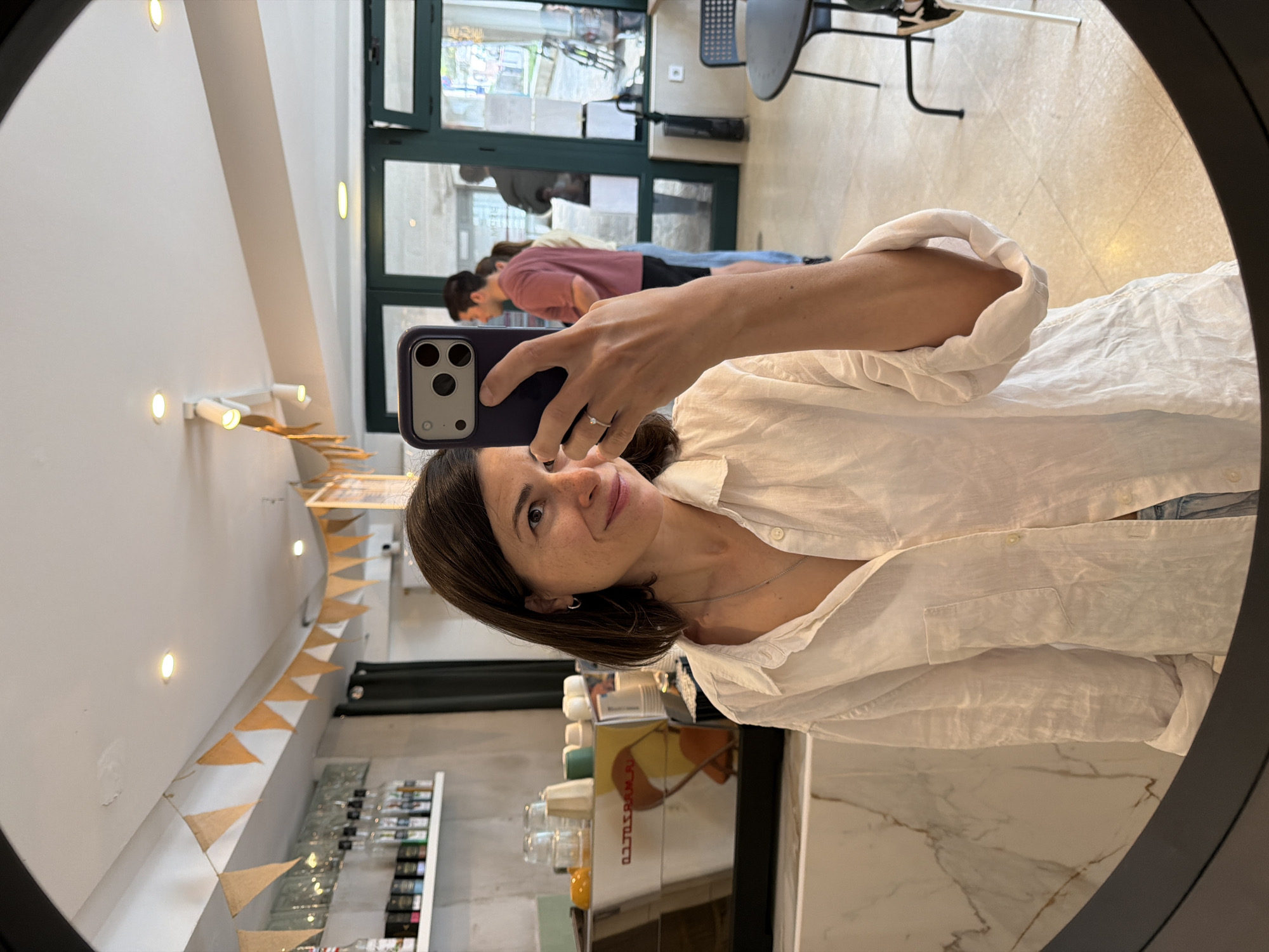 A selfie photograph shows a woman in a white shirt holding a smartphone, reflected in a round mirror. The background reveals a café or restaurant interior. Several people are visible behind her, including one wearing a pink shirt. In the background, there is a doorway or entrance. A display case with bottles, likely containing various liquids, and decorations along the ceiling are visible. A coffee machine and marble countertop are also within view. The location is Budva, Montenegro.