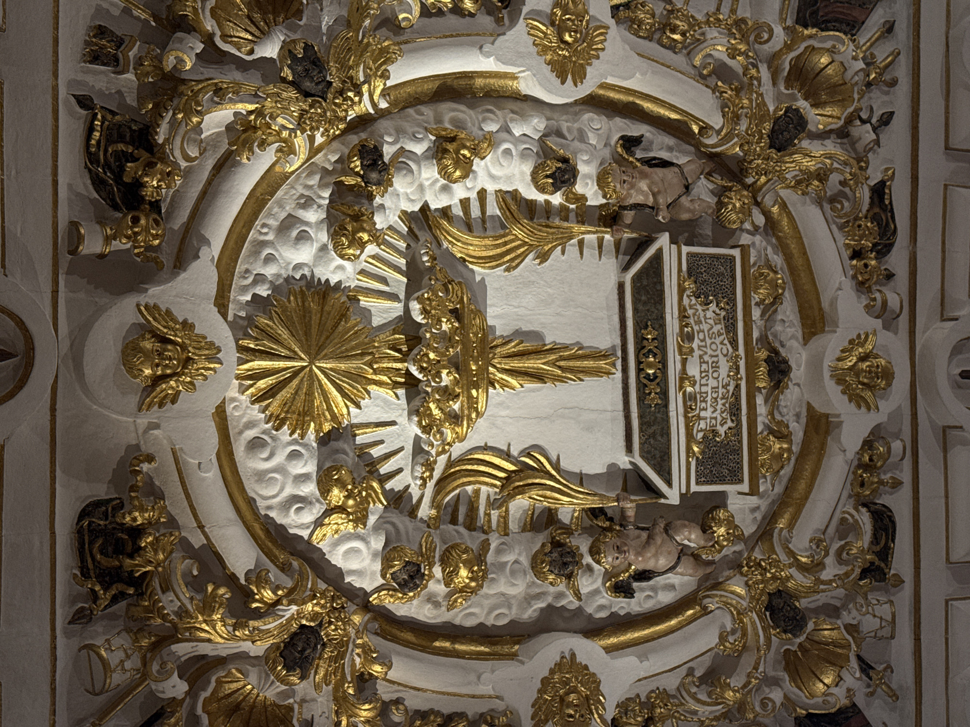 The image shows a detailed stucco and gold gilded relief sculpture depicting religious iconography. Central to the composition is a sarcophagus, possibly a reliquary, adorned with inscribed text in Latin. Above the sarcophagus, a gilded cross is crowned with a halo of rays. Flanking the cross are gilded palm fronds. The entire scene is framed by elaborate gold ornamentation, featuring swirling volutes, acanthus leaves, shells, and cherubic figures. Several cherubic heads with dark hair emerge from stylized clouds, some with gold wings. Two putti, one holding a gilded arrow, are positioned at either side of the sarcophagus, appearing to support or guard it. The sculpture is likely located within a church or religious building. The inscription on the sarcophagus appears to read "Erit sepulcri gloriosissimi", which translates to "It will be the tomb of the most glorious." Given the context of religious art and the inscription, this artwork is located in the Baroque Sacristy of the Cathedral of Santiago de Compostela, Spain. The sculpture is mounted on a white, textured wall.