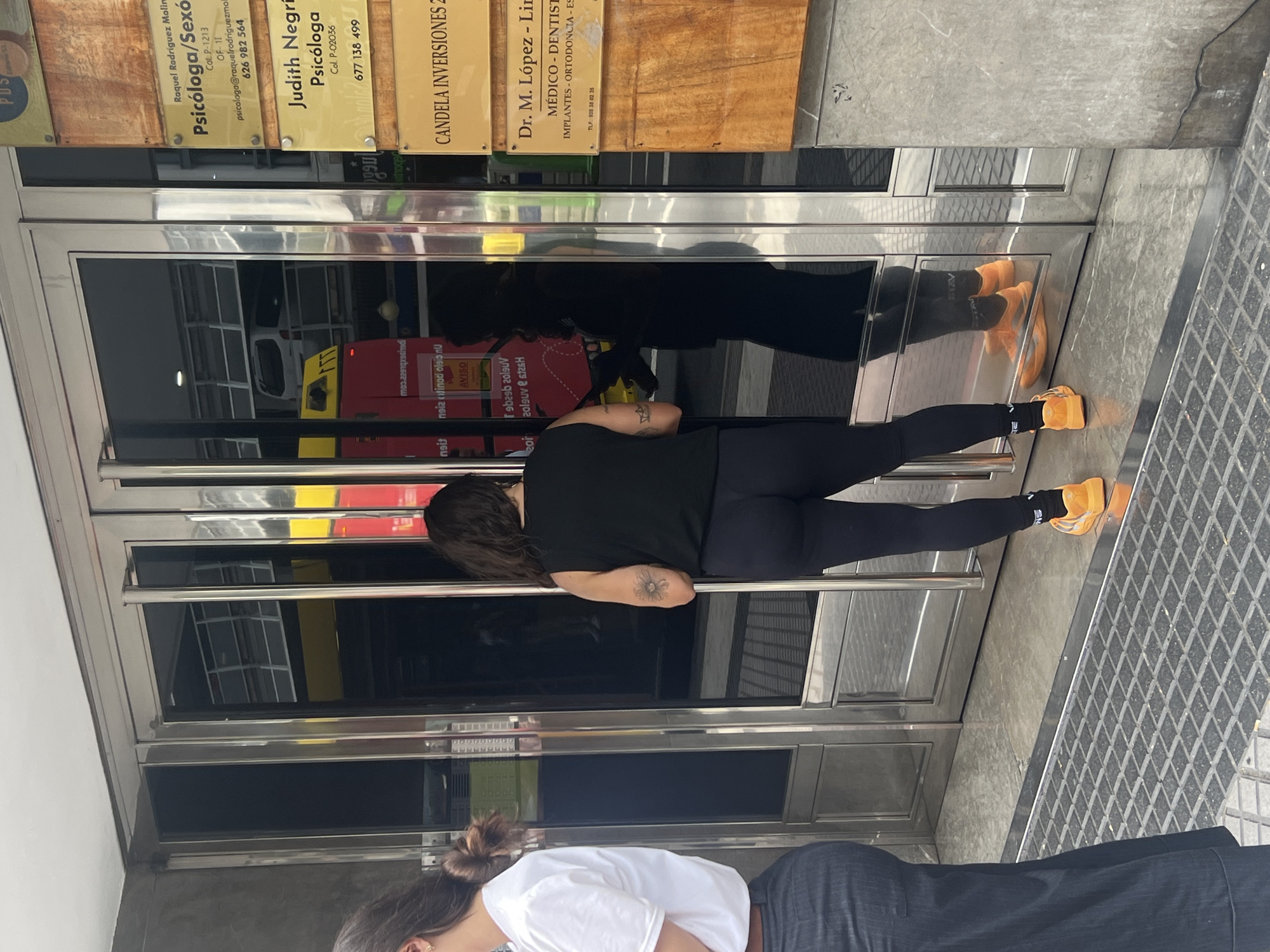 The image captures a bright daytime scene in what appears to be an urban setting, likely Las Palmas, Spain, depicting the entrance of a building. Two women are present, standing near tinted glass doors with prominent metal frames.
The woman in the foreground wears a black t-shirt, black leggings, and distinctive orange sneakers. She has dark hair and a visible tattoo on her upper arm. She is looking towards the glass doors, possibly at her reflection or waiting. Behind her, partially visible, is another woman wearing a white t-shirt and dark pants, with her hair tied up.
The glass doors reflect the surroundings, showing distorted images of the women, a white van or vehicle, and red and yellow signs, including one with the text "AVISO" and "comunicado."
Above the doors, several rectangular plaques provide information about businesses or professionals located within the building. Visible text on these plaques includes:
- "Raquel Rodríguez Molin / Psicóloga/Sexó / Col. P-1213 / OF-11 / psicologa@raquelrodriguezmoll / 626 982 564"
- "Judith Negri / Psicóloga / Col. P-02036 / 677 138 499"
- "CANDELA INVERSIONES 2" (partially visible)
- "Dr. M. López - Lin / MÉDICO - DENTIST / IMPLANTES - ORTODONCIA - ES / TLF: 920 38 02 35"
The ground is a mix of concrete and a dark, grated metal surface, typical of a sidewalk or building entrance area. The overall impression is that of people going about their day near commercial offices.