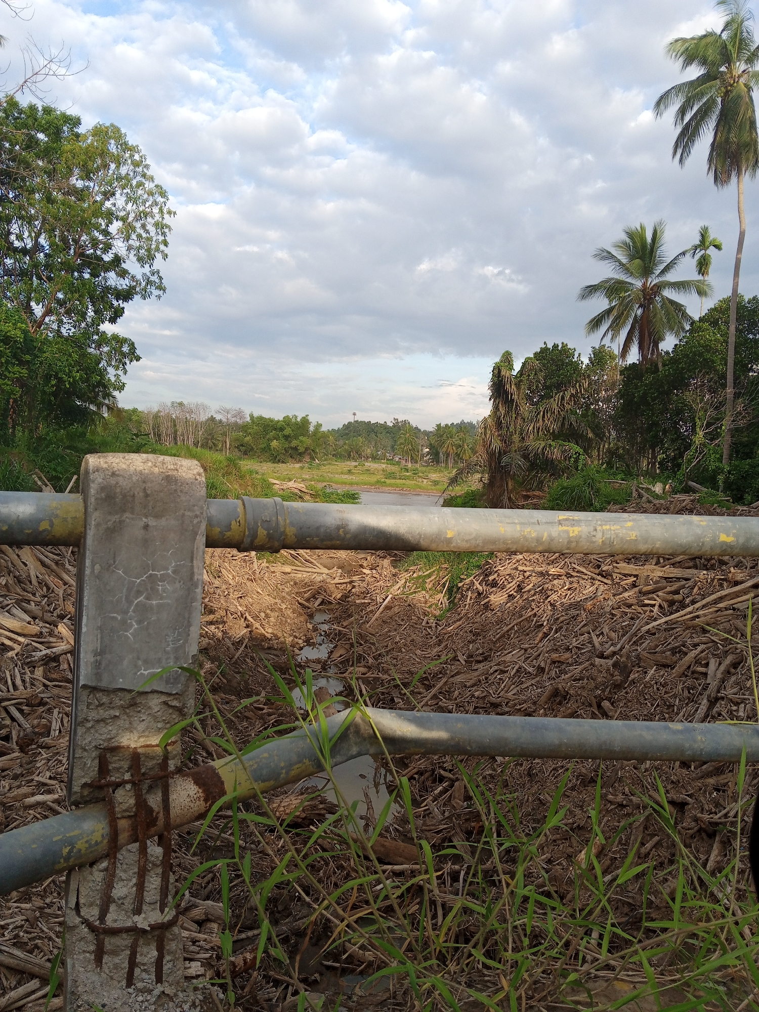 The image depicts a weathered landscape in Kruengbeukah, Indonesia. A dilapidated concrete and pipe barrier dominates the foreground, exhibiting rust and structural damage, with exposed rebar. Behind the barrier lies a terrain covered with wood debris and vegetation. A body of water is visible in the distance, bordered by dense tropical foliage and palm trees. The sky is overcast with clouds.