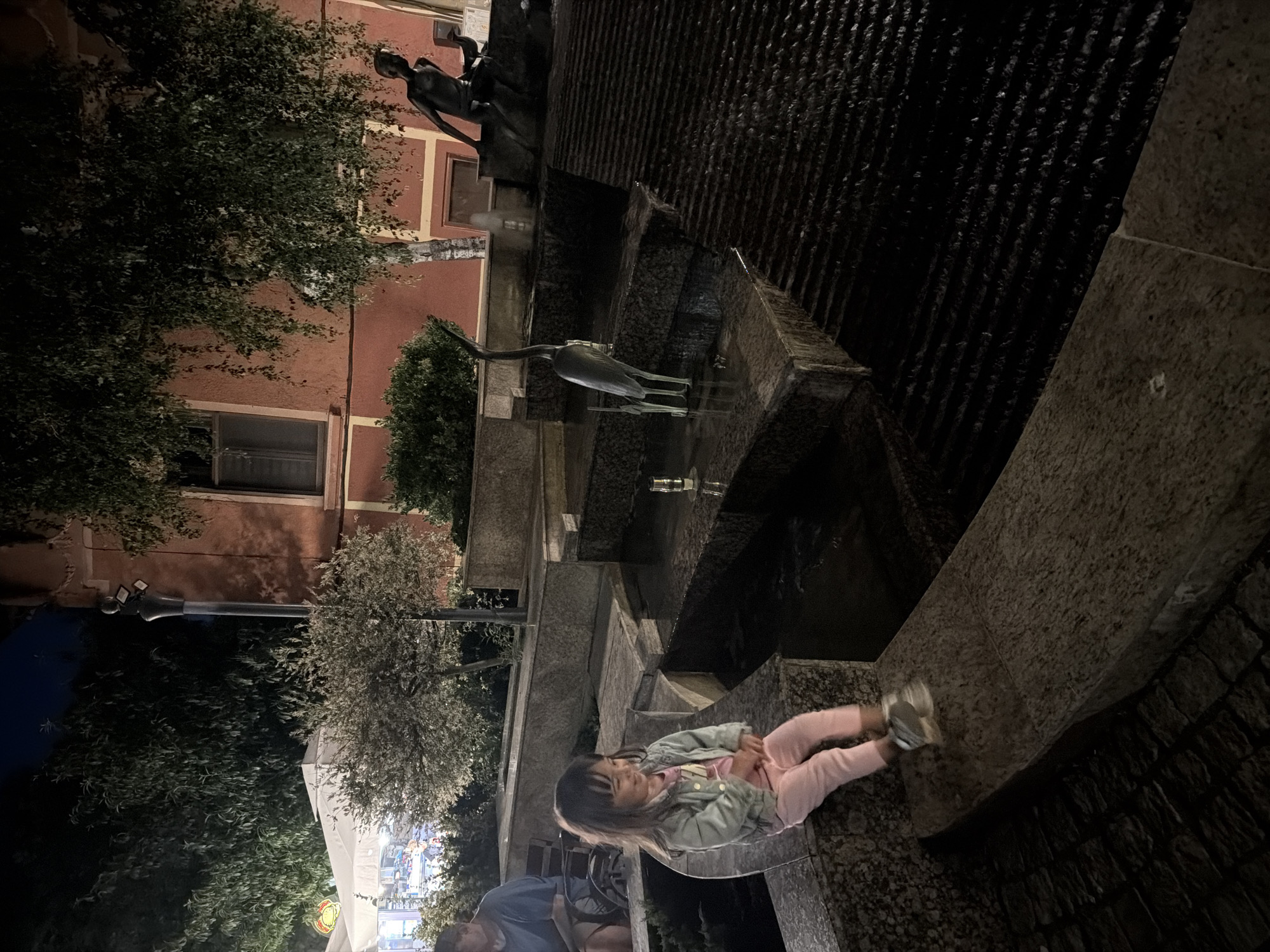 The image depicts a multi-tiered fountain at night in Burgas, Bulgaria. The fountain features stone steps with water flowing over them, creating a shimmering effect. A bronze sculpture of a heron stands on one of the tiers, and another bronze sculpture of a human figure is visible in the background. A young girl in pink pants and a light blue jacket is sitting on the edge of the fountain, looking to her right and smiling. She is wearing light-colored sneakers. In the background, a person is seated on a bench. The surrounding area is illuminated by artificial lights, highlighting the architectural features and vegetation. The buildings in the background have a reddish-orange facade. The overall scene conveys a calm and peaceful nighttime atmosphere in a public space.