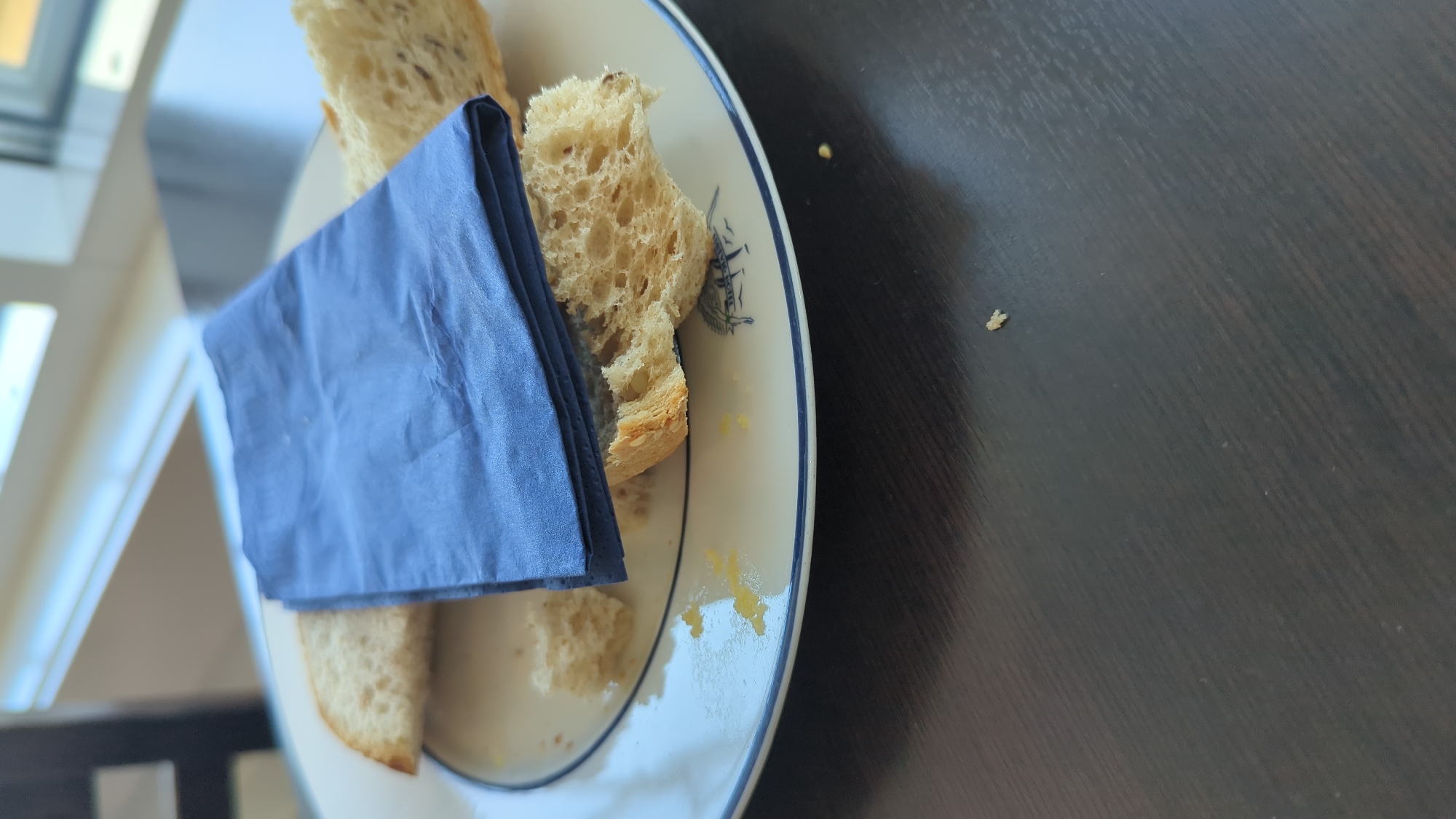 On a white plate with a blue rim, two slices of bread are placed. A blue cloth napkin is folded on top of the bread. There are crumbs on the plate and a small amount of yellow substance, possibly butter or jam, near the bread. The plate is on a dark brown wooden table. In the background, a window with a bright, blurred view of the outdoors is visible. The location is Larkollen, Norway.