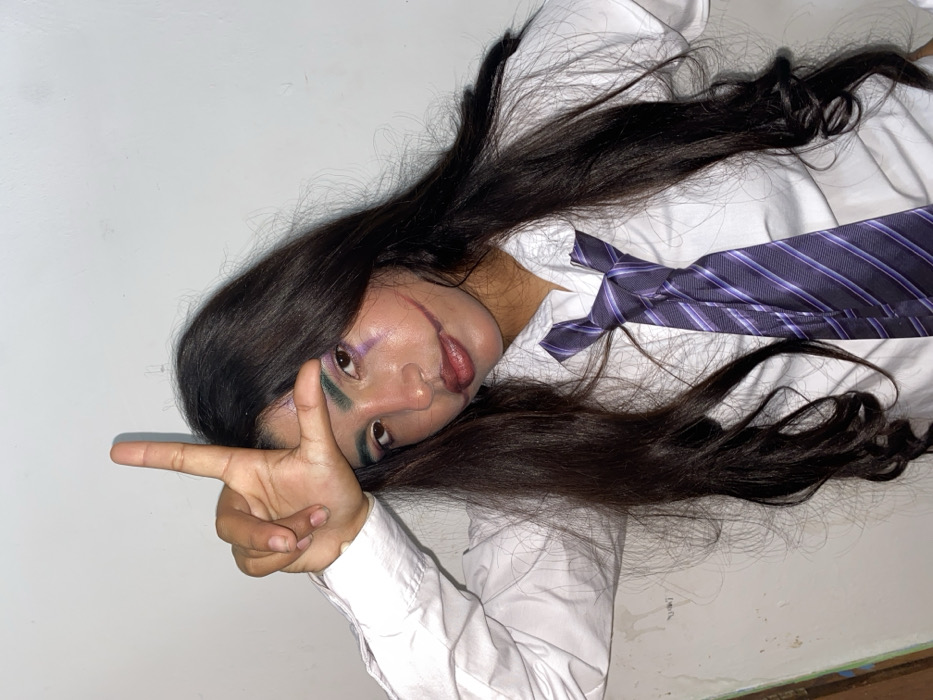 The image displays a head-and-shoulders portrait of a person, likely female, posed against a plain, light-colored background. The image is rotated approximately 90 degrees clockwise, making the subject appear to be lying down with their head to the left and their body extending to the right.
The person has long, dark brown hair that is spread out dramatically around their head and shoulders, covering parts of the white shirt they are wearing. Their face is turned towards the camera, and they are looking directly at the viewer. They are wearing elaborate makeup, featuring distinctive elements. Red or deep pink "scar" makeup extends from the corners of their mouth upwards onto their cheeks, creating the appearance of a "Glasgow smile" often associated with the Joker character. Their eyelids are adorned with vibrant makeup; the upper lid appears to have purple eyeshadow, while a striking teal or green color is smudged below the lower lash line. Their lips are painted with a glossy, possibly reddish-brown or deep pink color, blending into the scar effects.
The subject is wearing a white collared shirt with long sleeves, and a dark purple necktie with lighter diagonal stripes is loosely tied around their neck. The tie hangs casually over the front of the shirt.
Their right hand (from the viewer's perspective, which would be the left hand if the image were upright) is raised and positioned to form an "L" shape with the index finger extended horizontally to the left and the thumb extended vertically upwards. This gesture is commonly associated with the word "loser."
The background is a simple, undifferentiated white or off-white wall or surface, which helps to keep the focus entirely on the subject and their costume. There are some very faint, minor scuff marks or irregularities visible on the lower right edge of the background. The lighting is bright and even, highlighting the details of the makeup and costume.
The overall impression is that of a person portraying a character, likely for a costume event or photography session, with strong visual cues pointing towards a Joker-inspired theme due to the "Glasgow smile" makeup and possibly the "loser" hand gesture. No text is visible in the image.