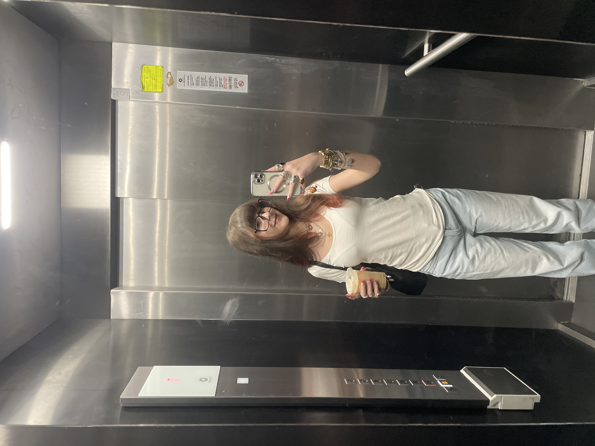 A woman is taking a selfie in an elevator. She is wearing glasses, a white shirt, and jeans. She is holding a drink in her right hand and her phone in her left hand. The elevator has metal walls and a control panel with buttons. The location is Raanana, Israel.