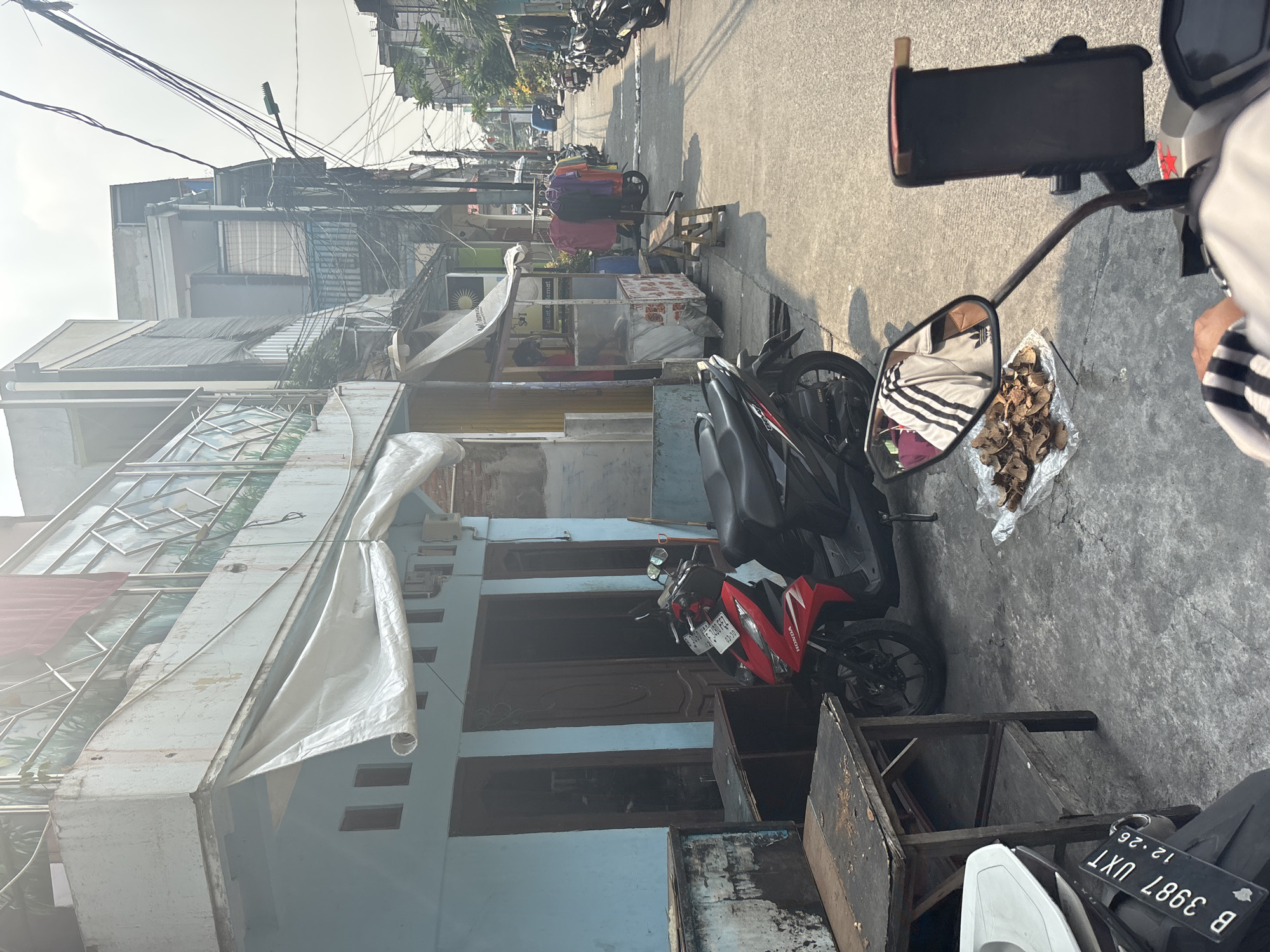 The image depicts a street scene in North Jakarta, Indonesia, likely a residential or commercial alleyway. In the foreground, a motorcycle with a red and black body is parked on a concrete ground. Next to the motorcycle, on a sheet of plastic, are dried leaves or food items. A wooden table is positioned to the left of the motorcycle. Behind the motorcycle is a light blue building with wooden doors and windows, covered partially by a white tarp. Across the street, a row of buildings with multiple stories can be seen, some with clothes hanging outside and small food stalls. Many other motorcycles are parked along the street, suggesting a common mode of transportation. The sky is bright, indicating daytime, and electrical wires are prominently strung across the scene. The overall impression is of a bustling urban environment with a focus on daily life and commerce.