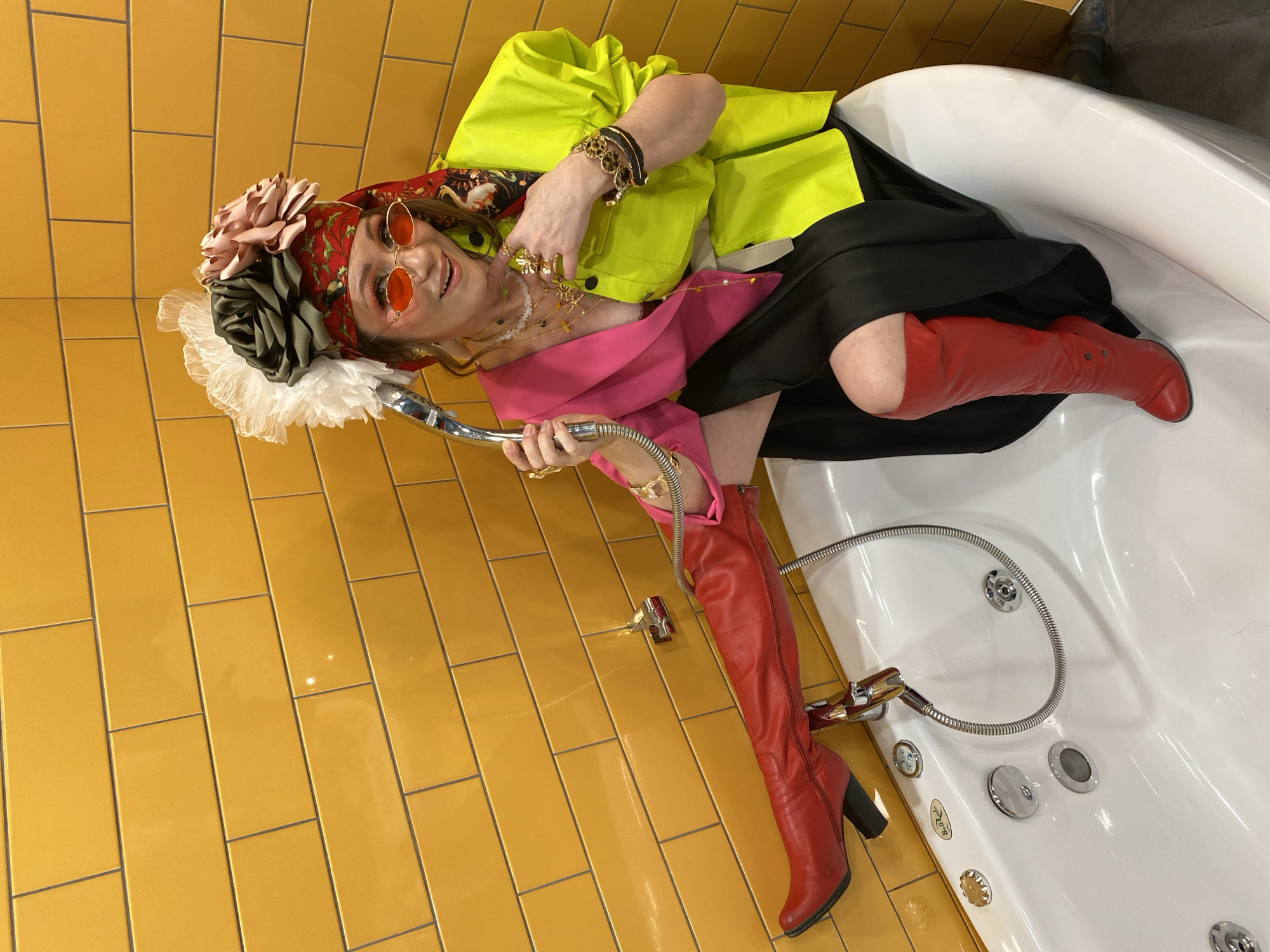 A woman is lying in a white bathtub with a silver handheld showerhead, in a room with yellow tile walls and floors. She is wearing red knee-high heeled boots, a black skirt, a pink shirt, a lime green jacket, and a red headscarf adorned with large artificial flowers. She also wears round-framed orange glasses and multiple bracelets and rings. The bathtub has multiple silver jets and fixtures. The scene is brightly lit.