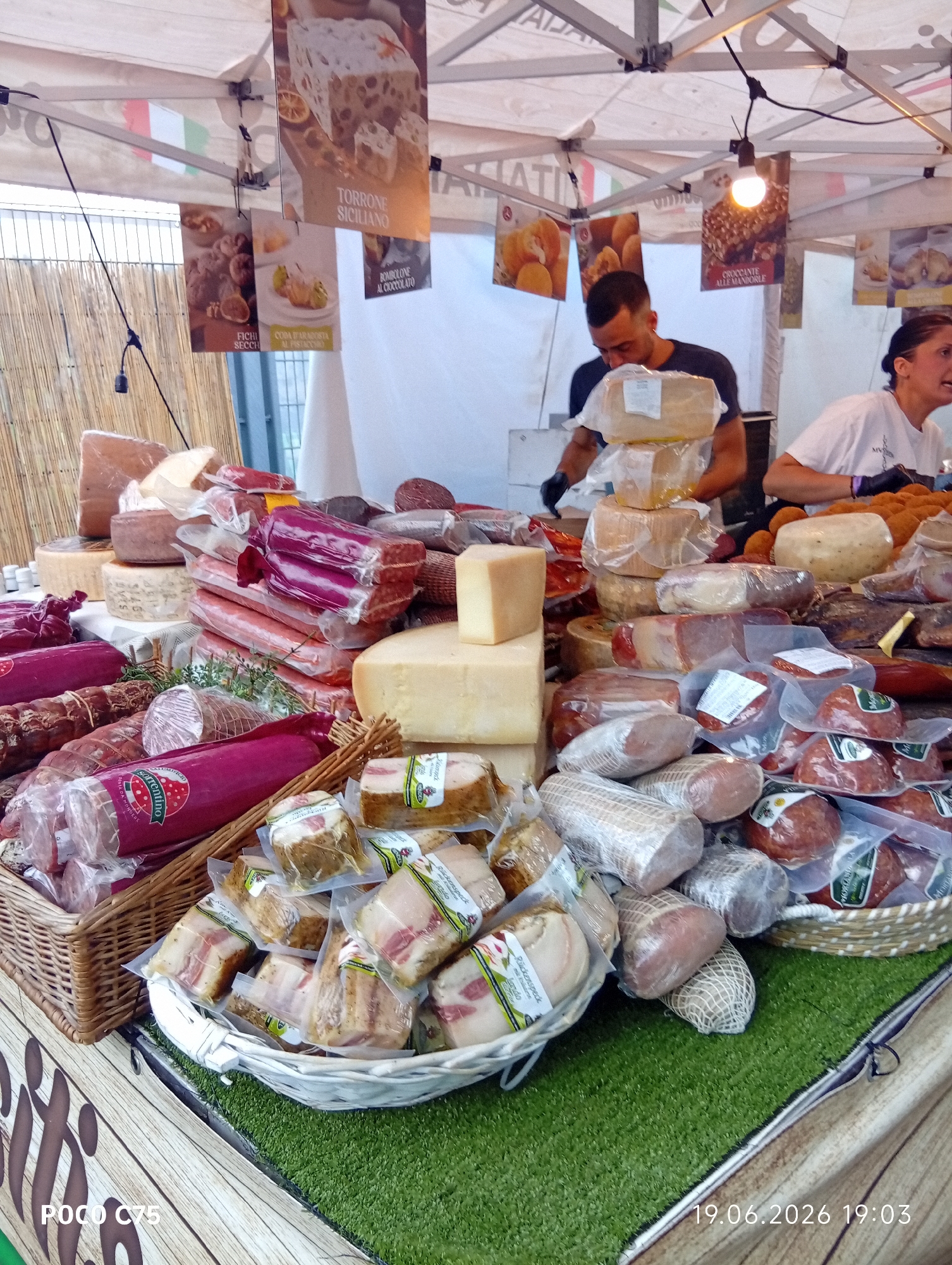 This media file shows a market stall laden with a variety of cured meats and cheeses. Two people, a man and a woman, are visible behind the counter, appearing to be vendors. The man is wearing a dark t-shirt and the woman a white t-shirt. They are engaged in serving customers or preparing items for sale. The stall is covered by a white canopy, and various signs with Italian text are hanging from it, advertising products like "TORRONE SICILIANO" (Sicilian Torrone), "FICHI SECCHI" (Dried Figs), "CODA BARADOSTA AL PISTACCHIO" (Pistachio Capo Staffa), "BOMBOLONE AL CIOCCOLATO" (Chocolate Doughnut), and "CROCCANTE ALLE MANDORLE" (Almond Brittle). The products themselves are prominently displayed, with many items, particularly the cured meats, wrapped in plastic packaging. Some are arranged in wicker baskets. The setting appears to be an outdoor market, possibly in a city, given the presence of a wooden fence-like structure in the background and the artificial green turf covering the display area. The timestamp at the bottom right indicates the image was taken on June 19, 2026, at 19:03. The general atmosphere suggests a busy marketplace at the end of the day.