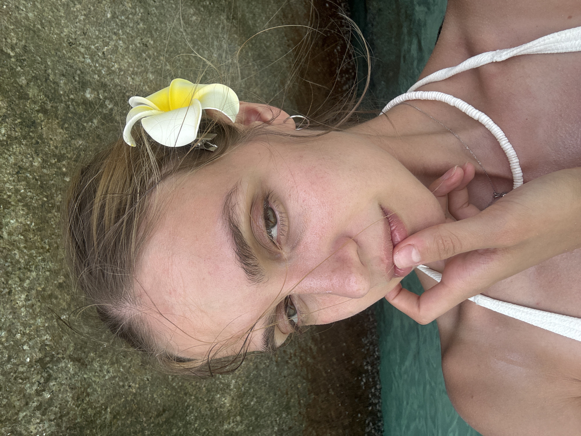 A young woman is pictured in what appears to be a coastal area, possibly in the city of Lam Kaen, Thailand, although no explicit landmarks confirm this. She is wearing a white top and has a white and yellow flower tucked behind her ear. Her hand is near her face, touching her lip. The background includes a rocky surface on one side and greenish water on the other. She is fair-skinned, and appears to be gazing slightly downward. A thin silver necklace is visible.