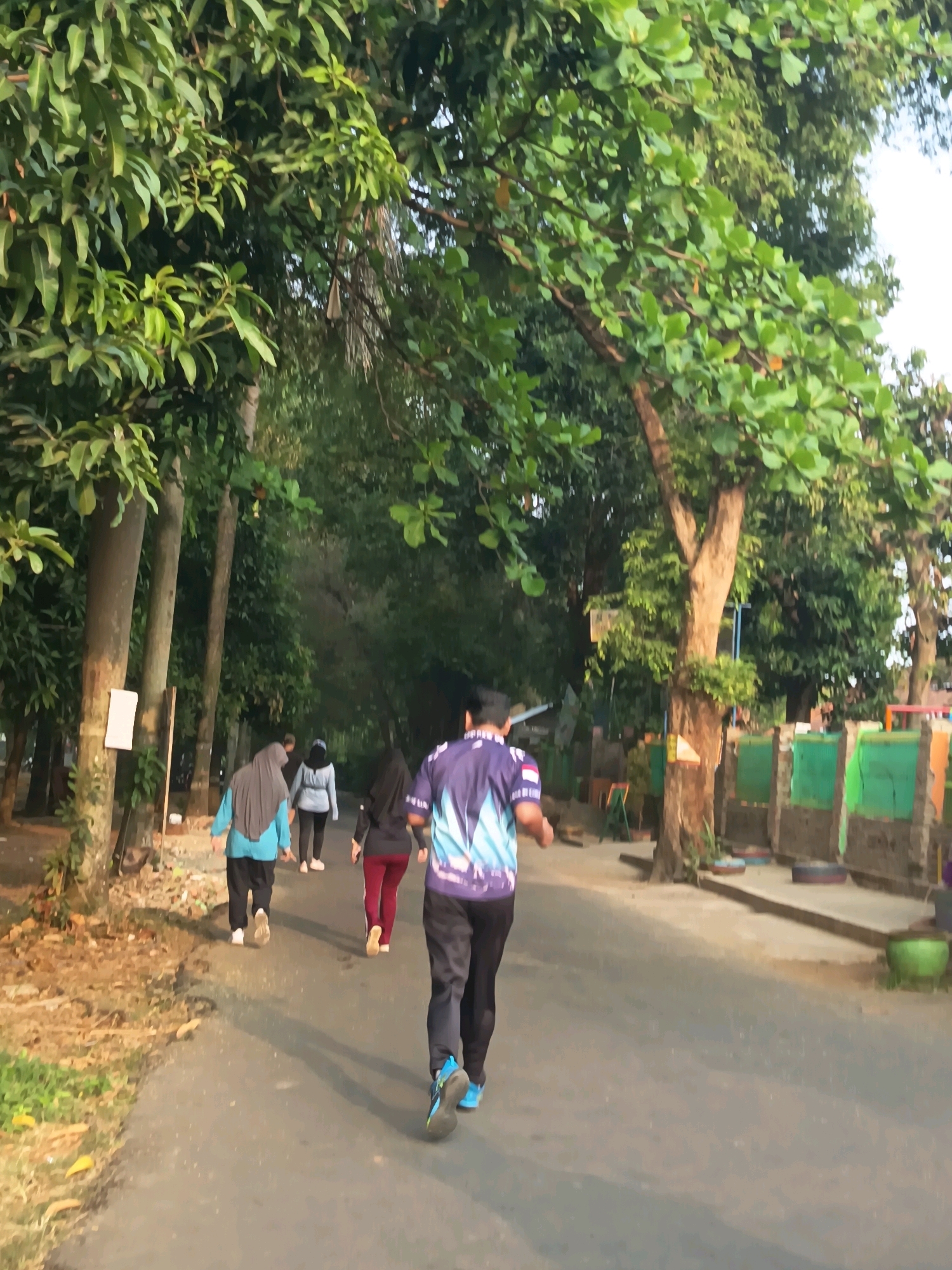 The image depicts an outdoor scene with several individuals engaged in physical activity. A person in a purple and blue athletic shirt and black pants is running on an asphalt path. Behind them, three other individuals, two women wearing hijabs and one in a black top and red pants, are walking. The environment is characterized by lush green trees lining the path, with dappled sunlight indicating it might be morning or late afternoon. The path appears to be in a park or a residential area with some fencing and structures visible in the background. The ground beside the path is covered in dirt and some scattered leaves. The overall impression is of a healthy outdoor activity taking place in a natural, green setting.