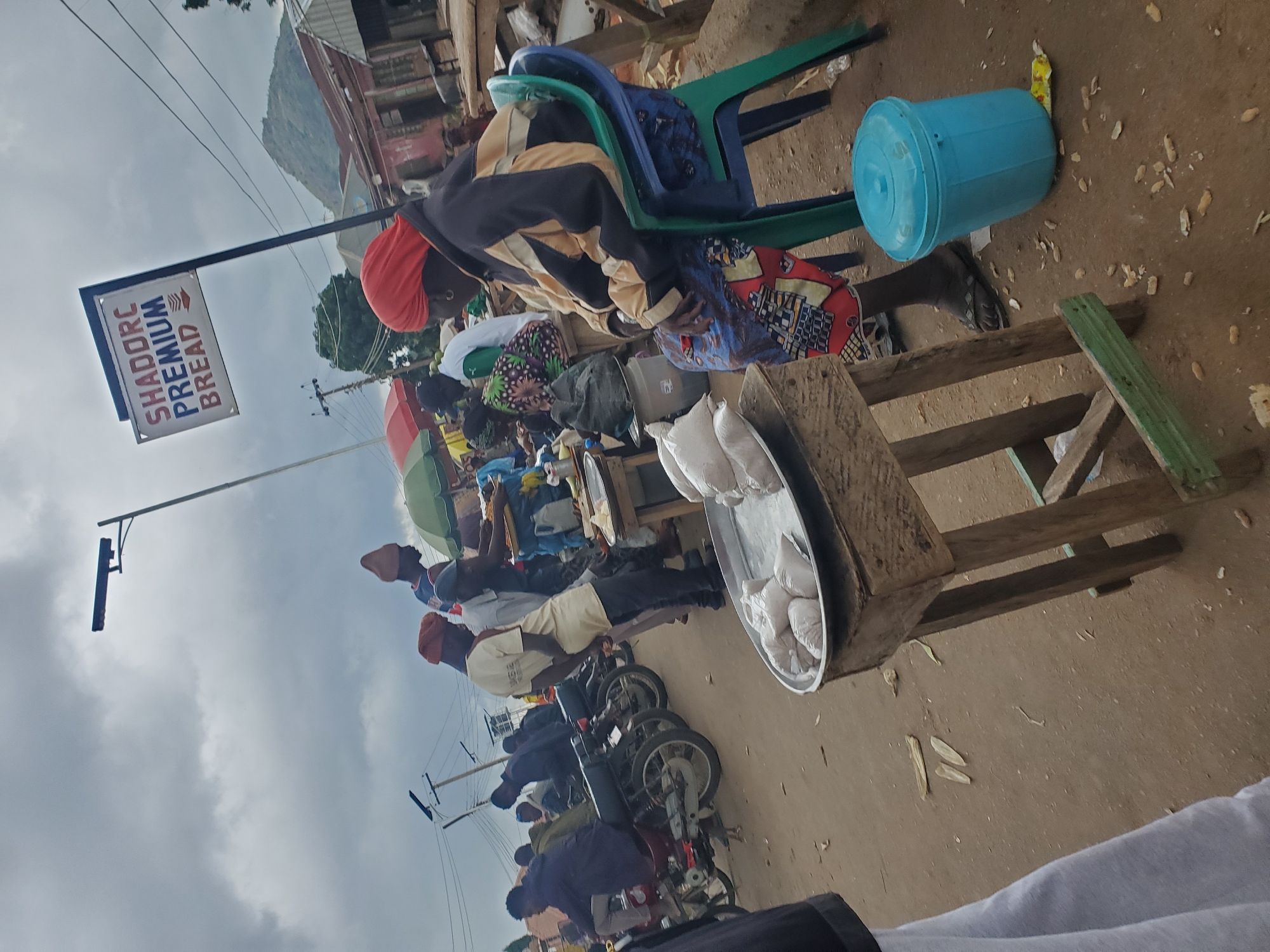 This image depicts a street scene in Gwong, Nigeria, likely a marketplace. In the foreground, a woman wearing a red headscarf and a colorful patterned skirt sits on a blue plastic chair. She is looking to her right. Beside her is a light blue plastic bin. In front of her, on a wooden stand, is a metal tray containing several white bags.
In the background, there are several people, some of whom appear to be vendors or customers. There are motorcycles parked in a line, suggesting transportation is a significant element of the scene. Overhead, a street lamp is visible, along with utility wires and poles. A sign with "SHADORC PREMIUM BREAD" written on it is mounted on a pole. Buildings with corrugated metal roofs are visible further in the background, and a distant, tree-covered hill or mountain is discernible under a cloudy sky. The ground is unpaved and dusty, with some debris scattered around. The overall impression is that of a bustling, everyday commercial activity in an outdoor setting.