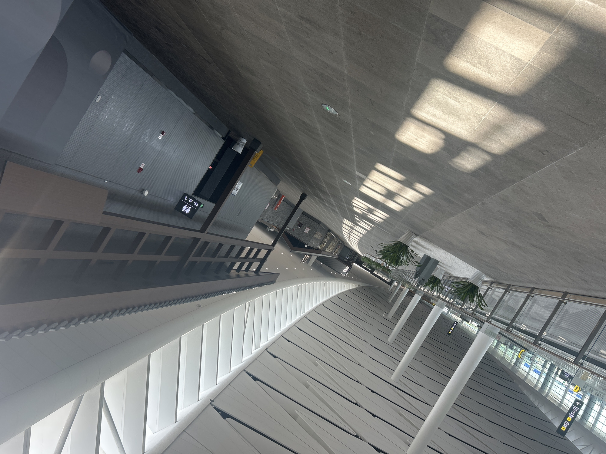 The image depicts a modern, spacious interior, likely an airport or a large public transit station, with a high, angled ceiling featuring a large skylight. The architecture is characterized by clean lines, white columns, and a grey tiled floor. Sunlight streams through the skylight, casting long, distinct rectangular shadows across the floor. Along the right side of the hallway, there are several potted plants. On the left side, a glass-enclosed walkway or waiting area is visible, with signage indicating directions and possibly gate numbers. Further down the hallway, a sign with male and female figures, presumably indicating a restroom, is mounted on a wall. The overall atmosphere is bright, clean, and contemporary. This location is within the city of Jianyang City, China.
