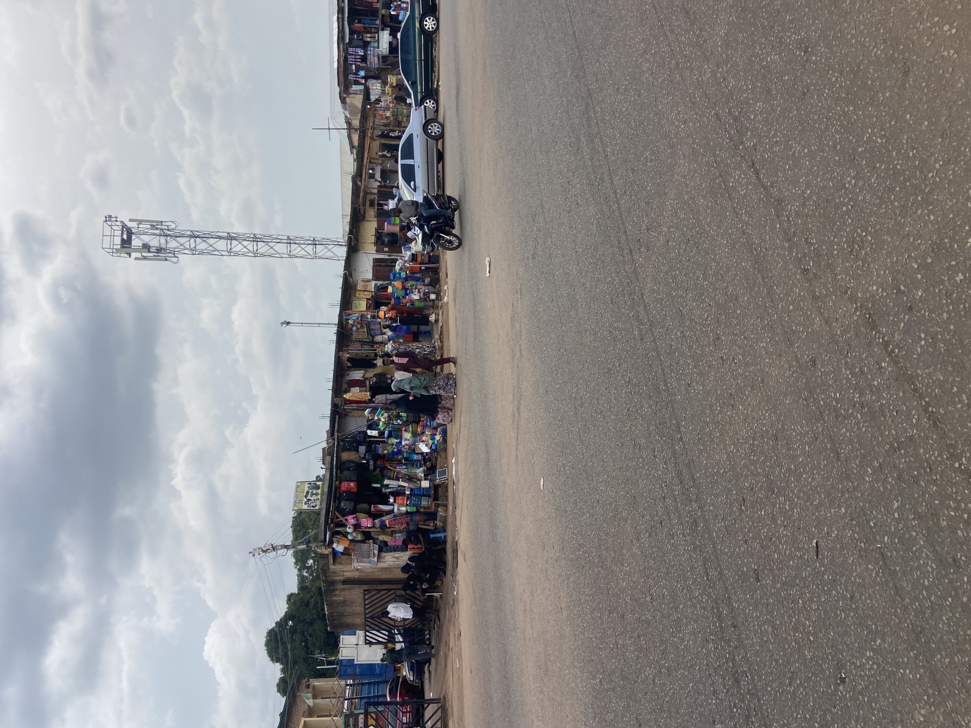 The image depicts a street market scene in Jos, Nigeria. A paved road runs along the right side of the frame, with a row of market stalls lining the left. The stalls are filled with a variety of goods, including clothing, bags, and what appear to be household items. Several people are visible, some tending to the stalls and others browsing the merchandise.
In the background, a large telecommunications tower stands tall, indicating a connection to modern infrastructure within the bustling market environment. A silver car and a dark green car are parked along the roadside near the market. A motorcycle is also present, with a person sitting on it.
The sky is overcast with clouds, suggesting it is likely daytime, possibly late morning or afternoon, and the weather appears to be cloudy. The overall atmosphere is one of everyday commerce and activity in a Nigerian urban setting. No discernible text is visible in the image.