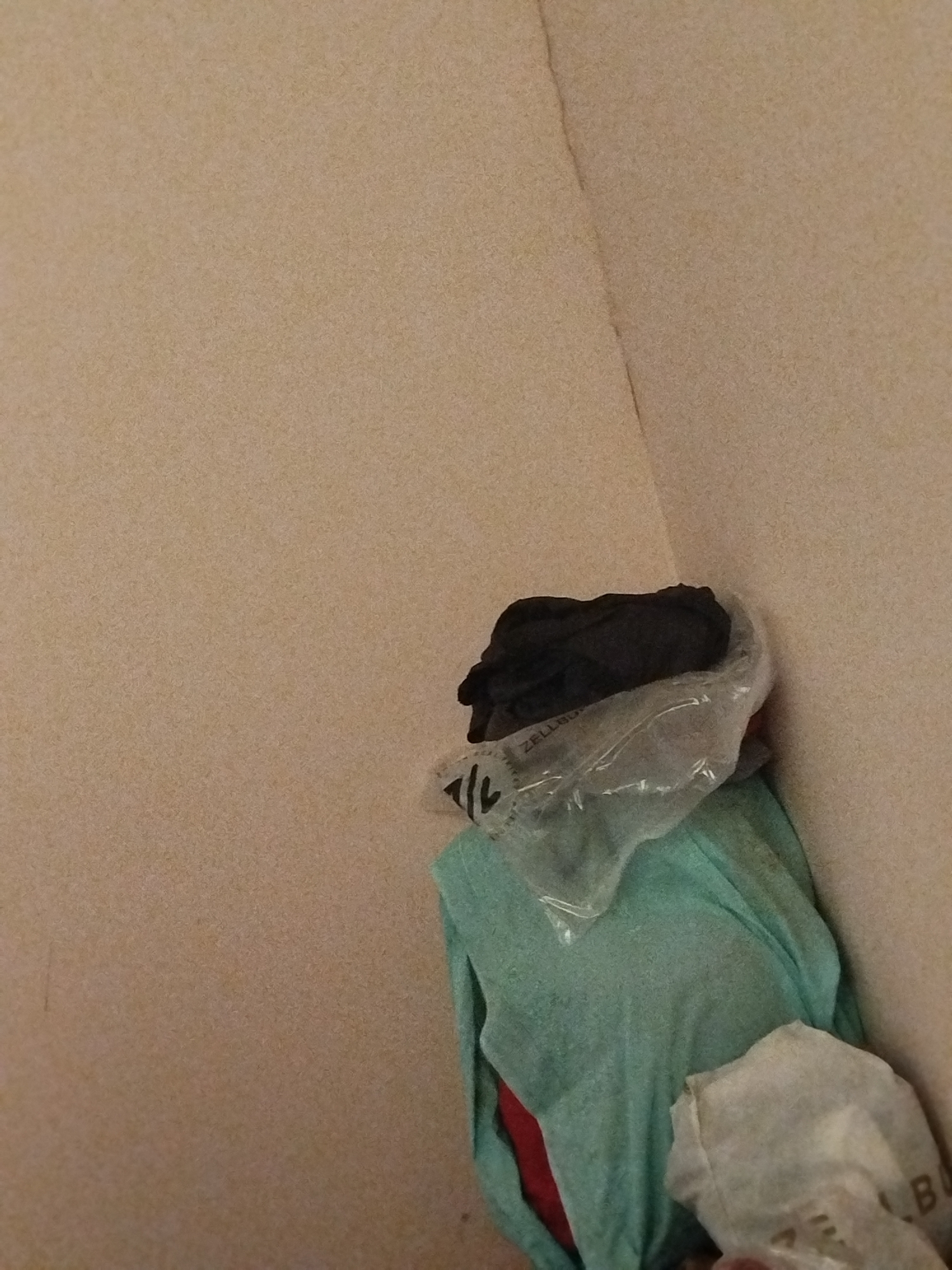 The image depicts a pile of clothing in the corner of a room. A light green garment is visible, with what appears to be a black garment on top, partially enclosed in clear plastic. Another white plastic bag containing items is partially visible in the foreground. The walls of the room are a neutral beige color. There is no indication of the city of Lassan, Pakistan, or any specific location within it.