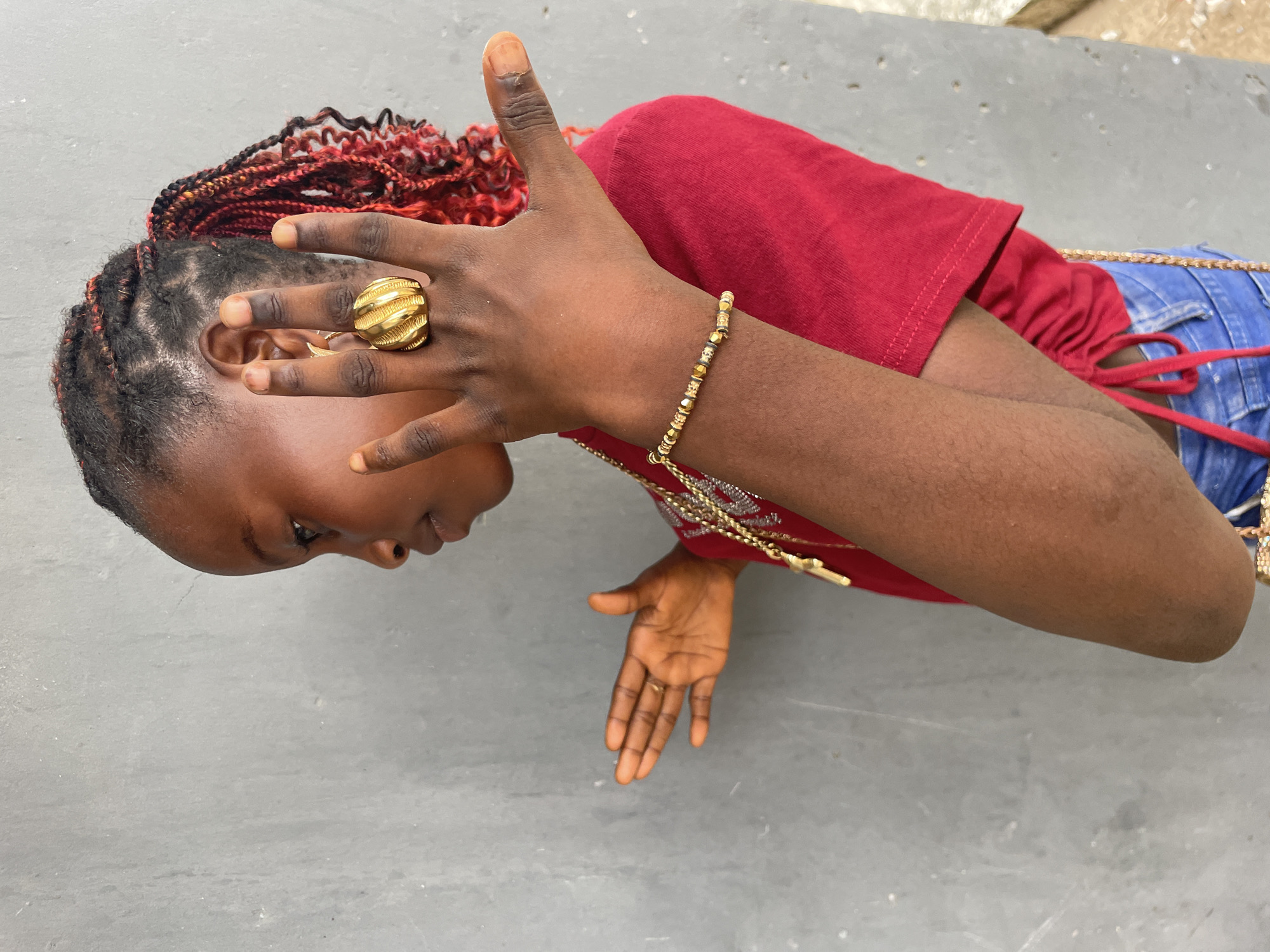 The image displays a young individual, likely female, from Badagry, Nigeria, positioned horizontally against a simple, light gray background. The subject has dark skin and her head is turned to her left, looking downwards, with a calm, focused expression. Her hair is intricately braided, featuring a striking mix of black, red, and orange hues.
She is wearing a short-sleeved red top and blue denim jeans, which are visible with a red tie-up detail. Her attire is accessorized with several gold-toned pieces: a large, textured gold ring with a ribbed design on the ring finger of her right hand, and a delicate gold-beaded bracelet on her right wrist. A long gold chain necklace with a decorative pendant is draped over her red top, and another gold chain, possibly part of a bag strap, rests on her jeans.
Her hands are central to the composition. Her right hand is raised near her face, with fingers slightly spread and thumb extended upwards. Her left hand is extended forward, palm open and fingers gently spread, suggesting a deliberate pose or gesture. The lighting is bright and even, indicating daylight. There is no visible text within the frame, and the minimalist background offers no specific location cues beyond the context of Badagry, Nigeria.