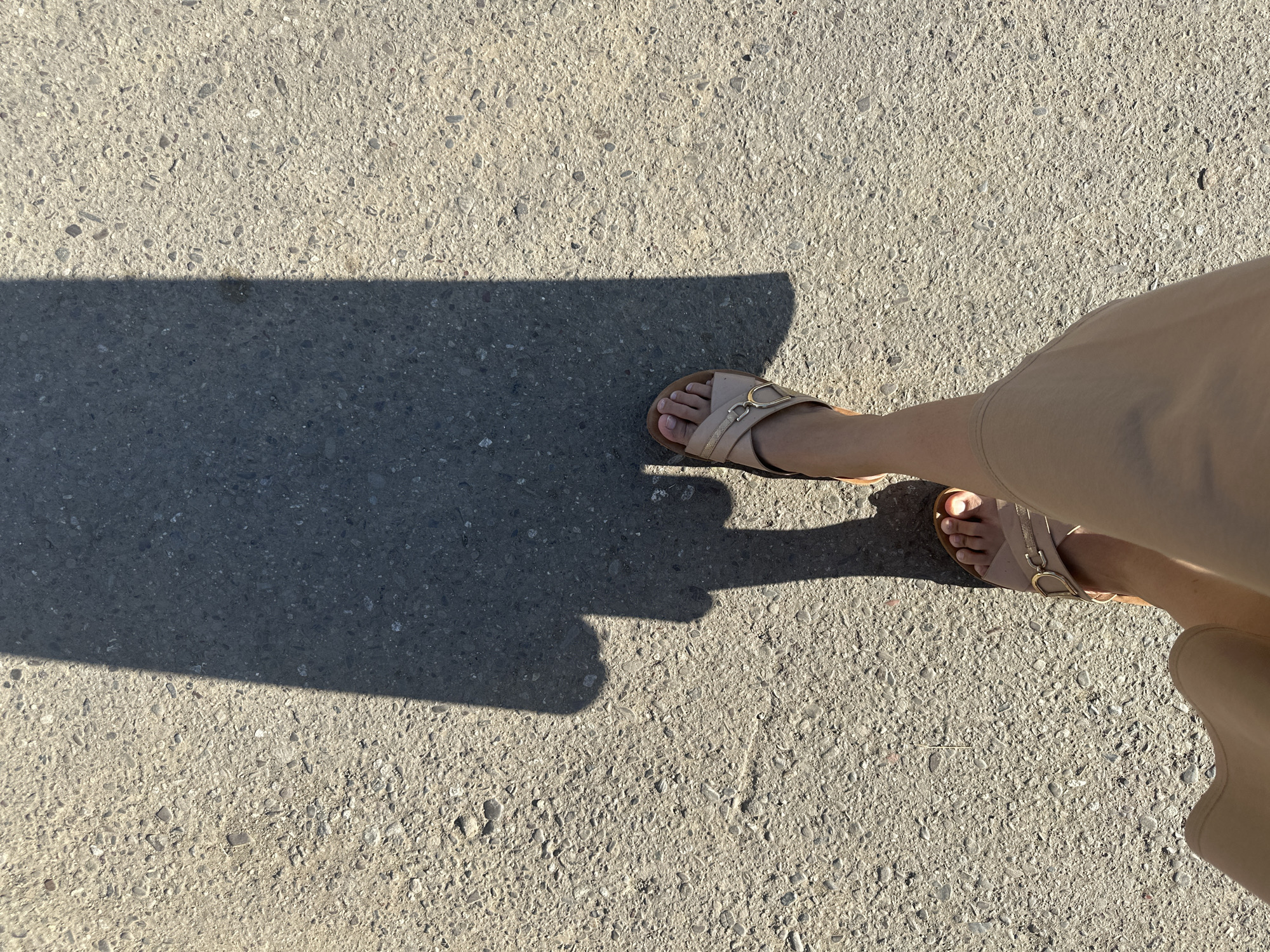An overhead shot on a concrete surface. The lower right portion of the image shows a tan-skinned person wearing tan slacks and tan sandals with gold buckles. The image includes the person's feet in the sandals. A dark shadow is cast across the left side of the concrete, obscuring detail in that area. The concrete surface is textured with small pebbles or aggregates. The location and context of this image are not discernible from the visual information provided.