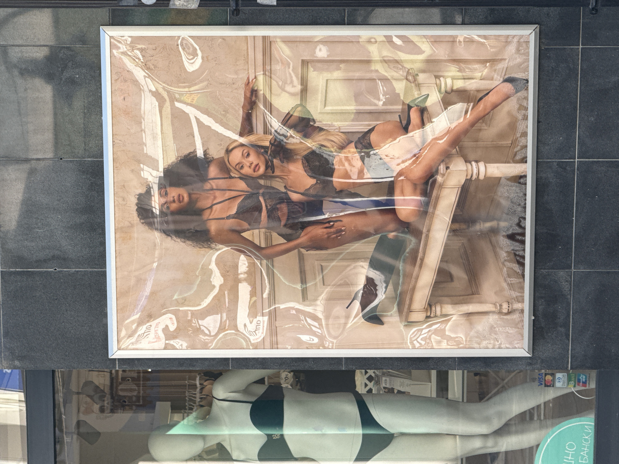 The image captures a display outside a retail store in Burgas, Bulgaria. A framed poster, depicting two women in lingerie, is placed above a mannequin wearing a bikini. The poster, partially covered by a transparent protective layer, features a woman with dark curly hair lying down next to a blonde woman, both posed in a suggestive manner. Below the poster, a mannequin is dressed in a dark bikini set. The scene is viewed from a slightly elevated perspective, suggesting the photographer was standing above the display. Payment options like Visa and Diners Club are visible on the storefront.