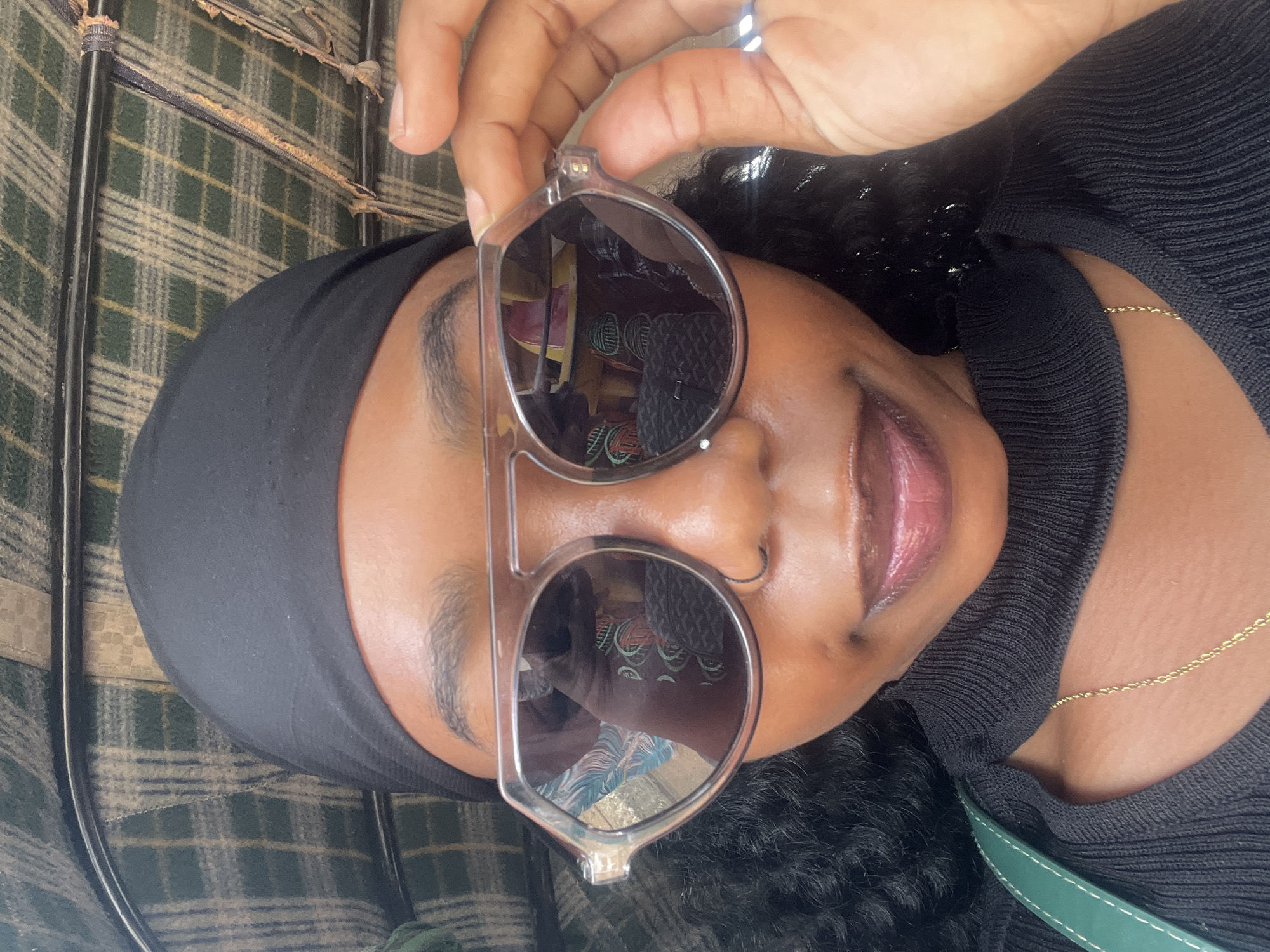 A close-up, selfie-style, medium shot captures a young Black woman from the chest up. She is inside a vehicle with a green and tan plaid headliner. The woman is wearing a black headscarf, dark sunglasses with a thick, transparent frame, and a black, high-necked top with a cut-out detail. A delicate gold chain necklace is visible at her collarbone, and a green strap from a bag is seen on her left shoulder. Her right hand is raised, holding the edge of her sunglasses. The overall lighting suggests it is daytime. The background, the plaid headliner of the vehicle, indicates she is likely within the city of Barakin Lerawi, Nigeria.