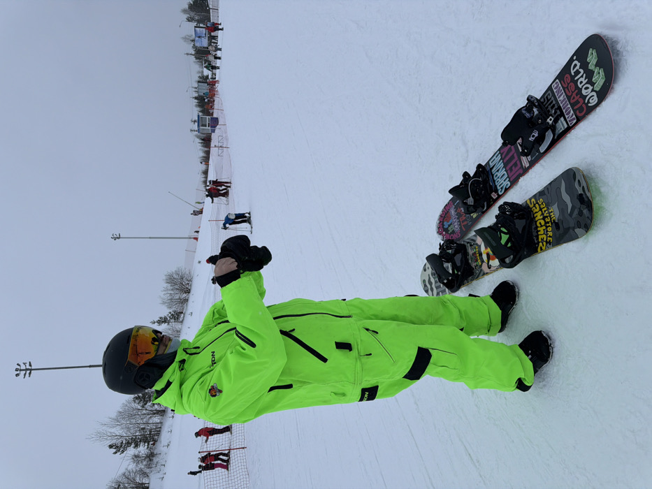 A person wearing a neon green ski suit and black helmet sits on a snowy ski slope with two snowboards nearby. One snowboard has a black, pink, and white design, while the other has a camouflage pattern. Other skiers and snowboarders are visible in the background, along with ski lift infrastructure and bare trees. The sky is overcast.