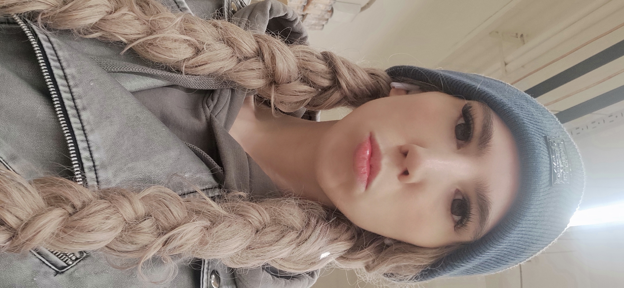 A fair-skinned young woman is shown in a close-up, tilted selfie. She has long, light blonde hair styled in two braids that frame her face. The woman wears a dark grey beanie and a grey jacket that has a zipper on its front. She has prominent eyelashes and full, pink lips. The background appears to be an interior setting, possibly a building in Chaikovsky, Russia, though no specific identifying landmarks or characteristics can be discerned from the image to confirm the exact location. The environment appears to be lit with artificial light, and the background is mostly out of focus.