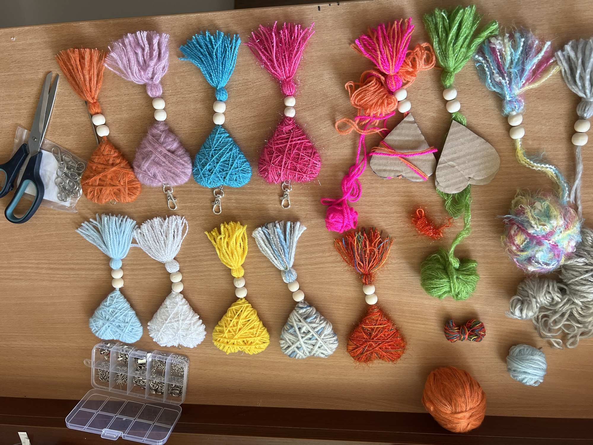 This image is a top-down view of a wooden surface, likely a table, displaying various craft supplies and unfinished projects. The scene is an indoor setting, with no visible indicators of its specific location within Białystok, Poland. The primary subjects are numerous handmade yarn tassels shaped like hearts, interspersed with balls of yarn in various colors including orange, light blue, grey, green, and multi-colored. Also present are a small plastic organizer box filled with metal findings such as lobster clasps and jump rings, and a pair of scissors. The tassels appear to be in various stages of completion, with some featuring wooden beads. The lighting suggests it is daytime.