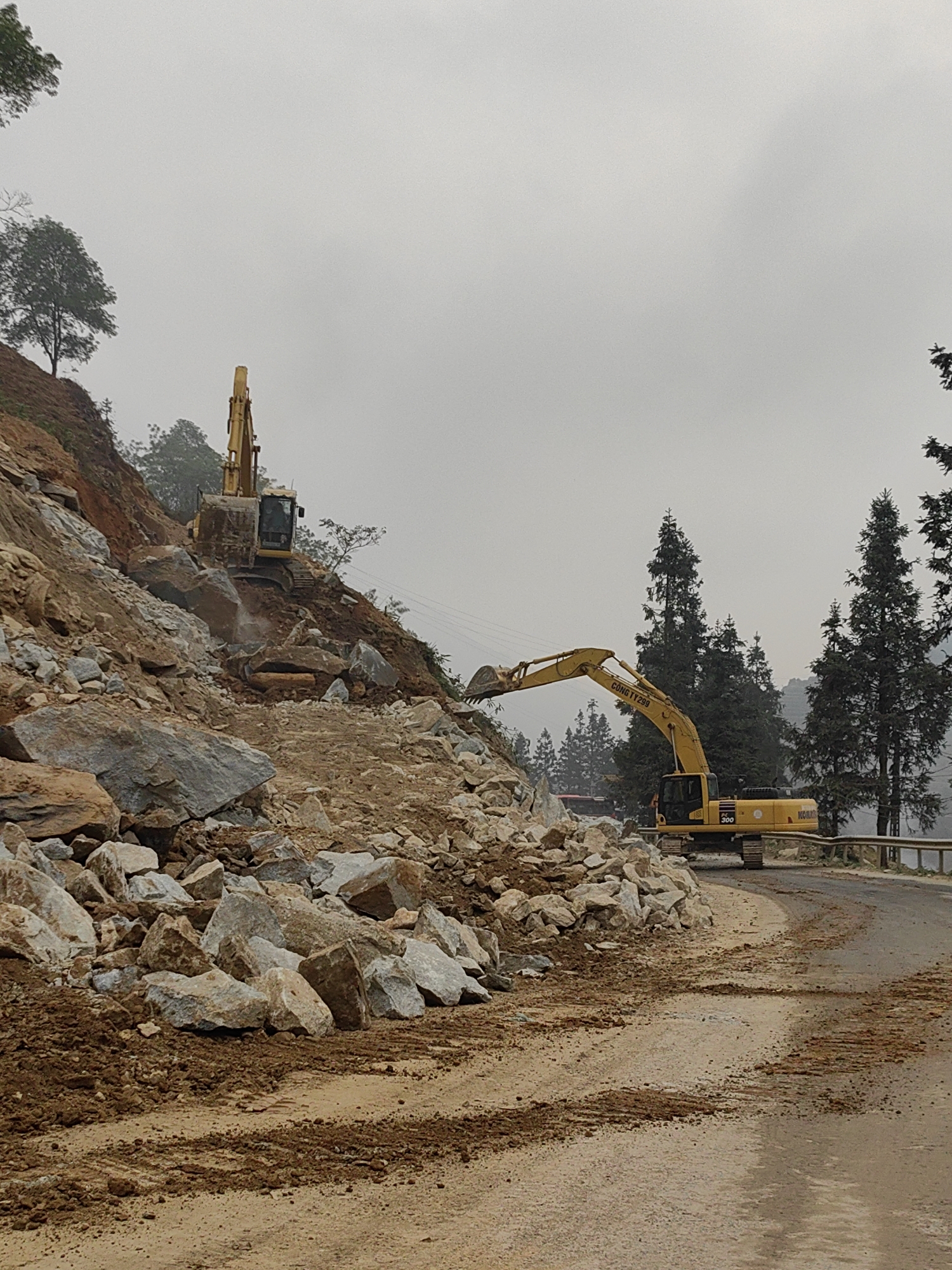 The image displays a road clearing operation in Trung Chải, Vietnam, following a substantial rockfall or landslide. A curved, paved road, partially covered with brown soil, mud, and light-colored rock debris, diagonally traverses the lower half of the frame. Two yellow hydraulic excavators are present. One excavator is positioned high on a steep, brown earth and rock-strewn hillside to the left, its bucket positioned amidst a large pile of fragmented rocks. A second, larger yellow Komatsu PC 300 excavator, marked "CÔNG TY 299", is located on the paved road in the middle-ground, with its arm extended towards a substantial rock pile encroaching on the left side of the road. Dense coniferous trees line the right side of the road and are visible in the background, partially obscured by an overcast, hazy sky. Utility lines are visible in the upper left background. A metal guardrail borders the right edge of the road further down the curve. No people are visible.