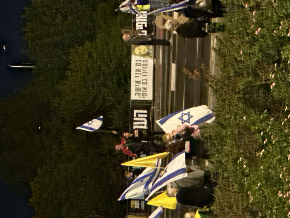 The image displays a nighttime outdoor scene in Mishmar Hasharon, Israel, capturing a gathering of approximately 15 to 20 individuals alongside a road. The foreground is filled with dense green foliage interspersed with small light-colored flowers. In the mid-ground, numerous people are visible, many holding Israeli flags, which are white with a blue Star of David. One individual is holding a plain yellow flag. A large banner with Hebrew text and a photographic portrait of a woman is prominently displayed behind the group; the Hebrew translates to "I am also a woman. Bring me back too." A man wearing a red top is seen holding an Israeli flag aloft in the upper-center, and a child holding an Israeli flag is present in the lower-center. A paved road surface is partially visible behind the group. The background consists of dense green trees, and a streetlight pole with an illuminated fixture is discernible in the upper-right. The entire scene is illuminated by artificial light sources.