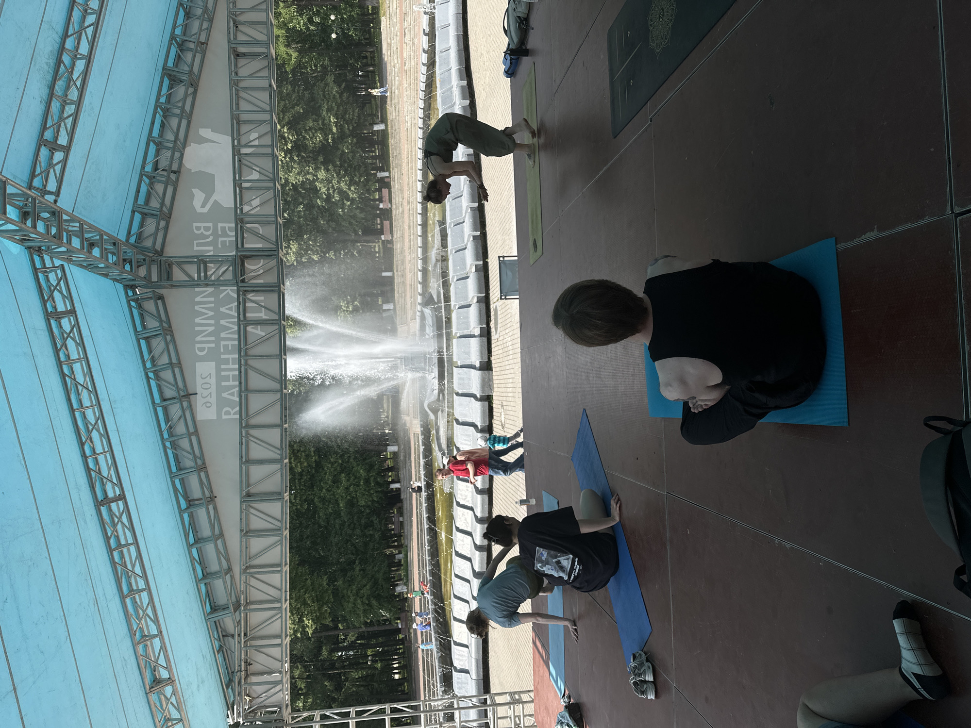 The image depicts an outdoor yoga session taking place on a raised wooden stage in front of a large fountain and tiered seating area, likely in a park or public square in Vladimir, Russia. A bright blue tarp, supported by a metal truss structure, forms a canopy over the stage, casting a shadow.
In the foreground, three individuals are engaged in yoga on mats. One person, wearing a black tank top, is seated with their legs crossed on a blue yoga mat. To their left, another person in a blue t-shirt is in a low plank or tabletop position on a blue mat, with a pair of grey sneakers and a dark bag nearby. Further to the right, a third person, wearing a green jumpsuit, is standing on a green mat, mid-movement, with their hands held out.
In the mid-ground, behind the stage, a fountain is actively spraying water. Several people, including adults and children, are visible near the fountain and seating area. Two men in casual clothing and a child in a turquoise outfit are interacting near the fountain's edge. More figures are scattered throughout the background, including people walking and sitting. The environment appears to be a sunny day with lush green trees framing the scene. A banner with Cyrillic text and a silhouette of a person in a yoga pose is visible behind the truss structure.