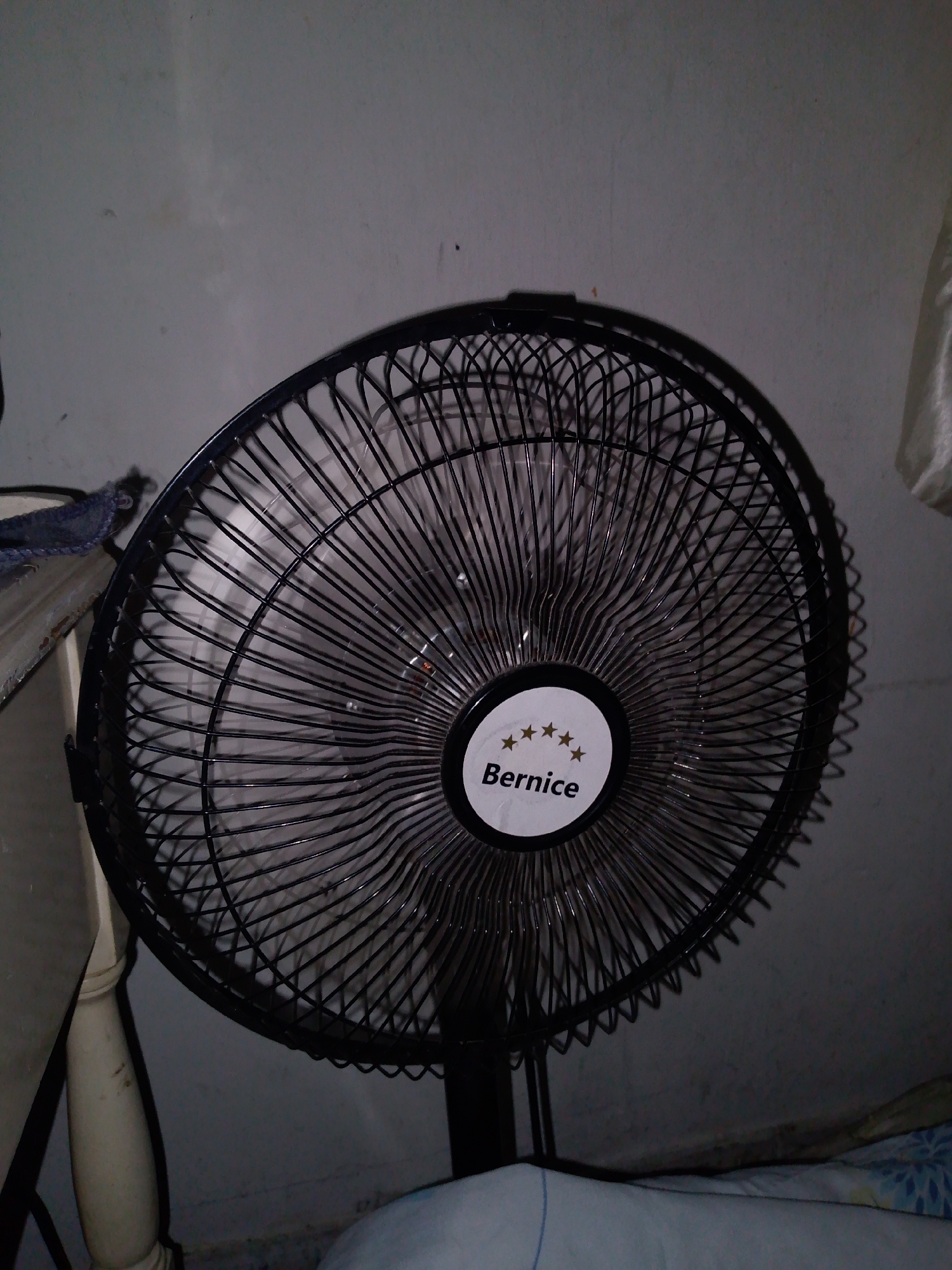 A black Bernice brand oscillating fan is positioned in a dimly lit indoor space. The fan is characterized by its circular protective metal cage with thin, closely spaced wires, and a white central logo displaying "Bernice" beneath five gold stars. The fan stands on a black pole-like base. Behind the fan is a light gray wall, with portions of furniture and bedding visible on either side of the fan. The setting appears to be a room, possibly a bedroom, inside a residence. There is no evidence to connect this image with the city of Caracas, Venezuela.