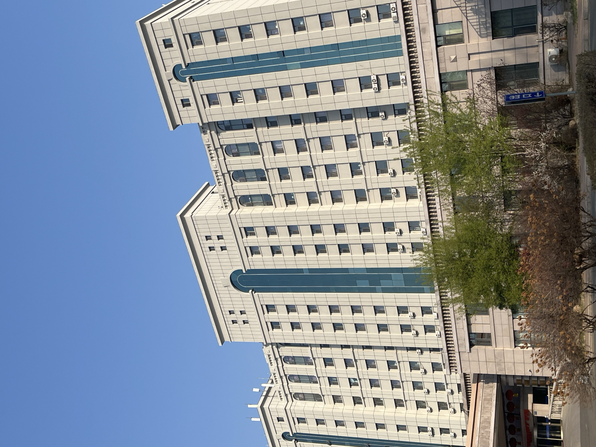 The image displays a modern, multi-story building under a clear, bright blue sky. The building features a light-colored facade, likely beige or light grey, with numerous rectangular windows arranged in a grid pattern. Horizontal bands of reflective blue material, possibly glass, run vertically up the building's exterior, punctuated by decorative semi-circular arches. Numerous small, white air conditioning units are mounted below many windows across different floors.
To the right of the building, at ground level, there are several trees, some with vibrant green leaves suggesting spring or early summer, and others with dry, brown foliage. A blue street sign is visible among the trees and road, displaying the Chinese characters "路口" (lùkǒu), which translates to "intersection," along with a downward-pointing arrow. A paved area, likely a road or sidewalk, is visible below the trees. The bright, cloudless sky indicates it is daytime with fair weather. No explicit activity or people are depicted in the scene, which is set in an urban environment in Changchun City, China.