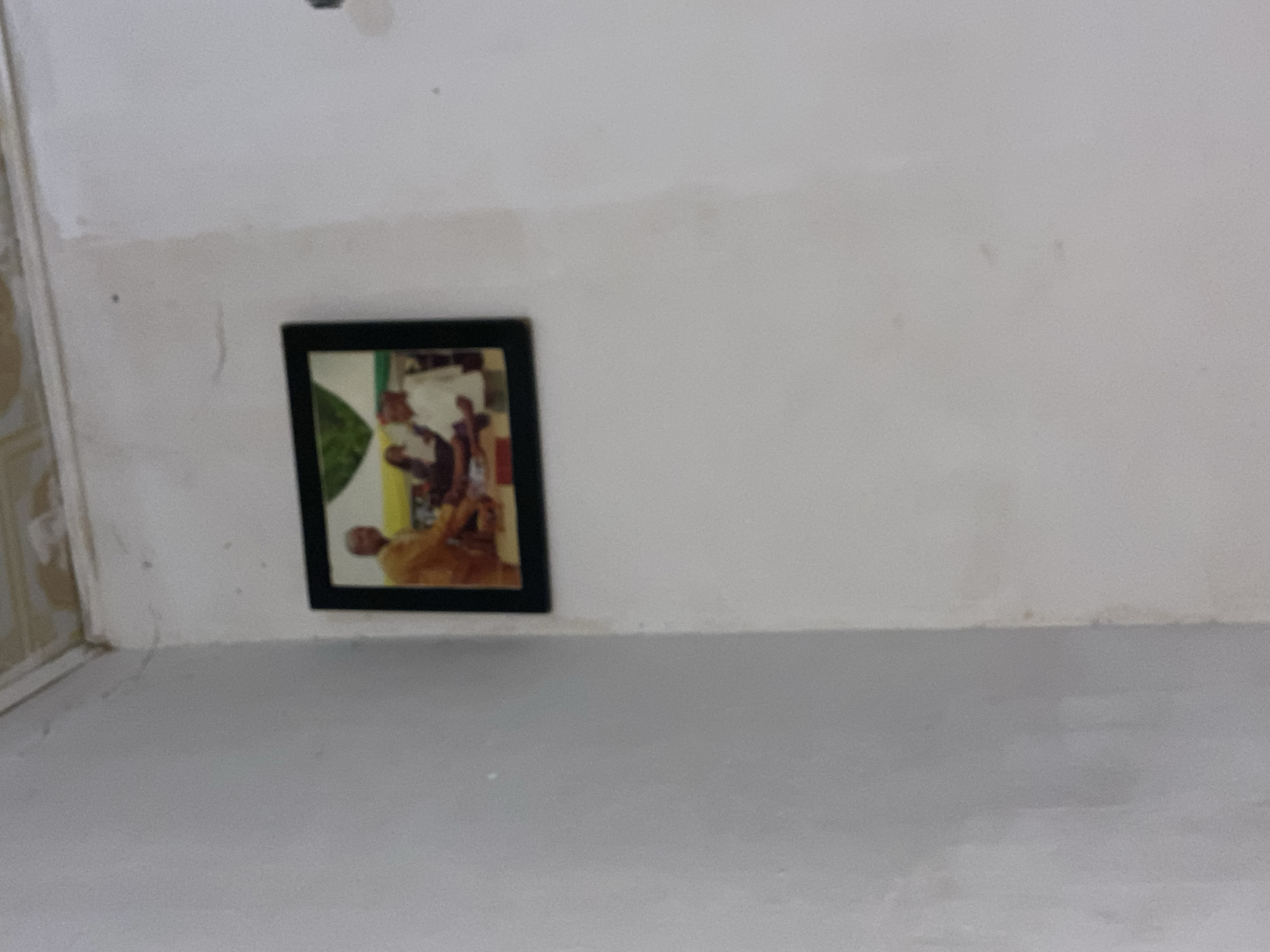 A framed photograph hangs on a white wall in a room. The photograph depicts two men in a handshake. One man is wearing a yellow traditional outfit, and the other is in a white shirt. The background of the photograph shows a blurred green and yellow scene, possibly indicating an outdoor event or a backdrop. The wall itself appears to be slightly aged, with some minor marks and a crack near the ceiling. The ceiling, visible in the upper part of the image, also shows signs of wear. The lighting in the room is ambient, casting a soft glow on the scene. This photograph is likely a personal memento, possibly of a significant event or a meeting with an important individual. The overall setting suggests a common indoor space, perhaps a home or an office, in Jalingo, Nigeria.