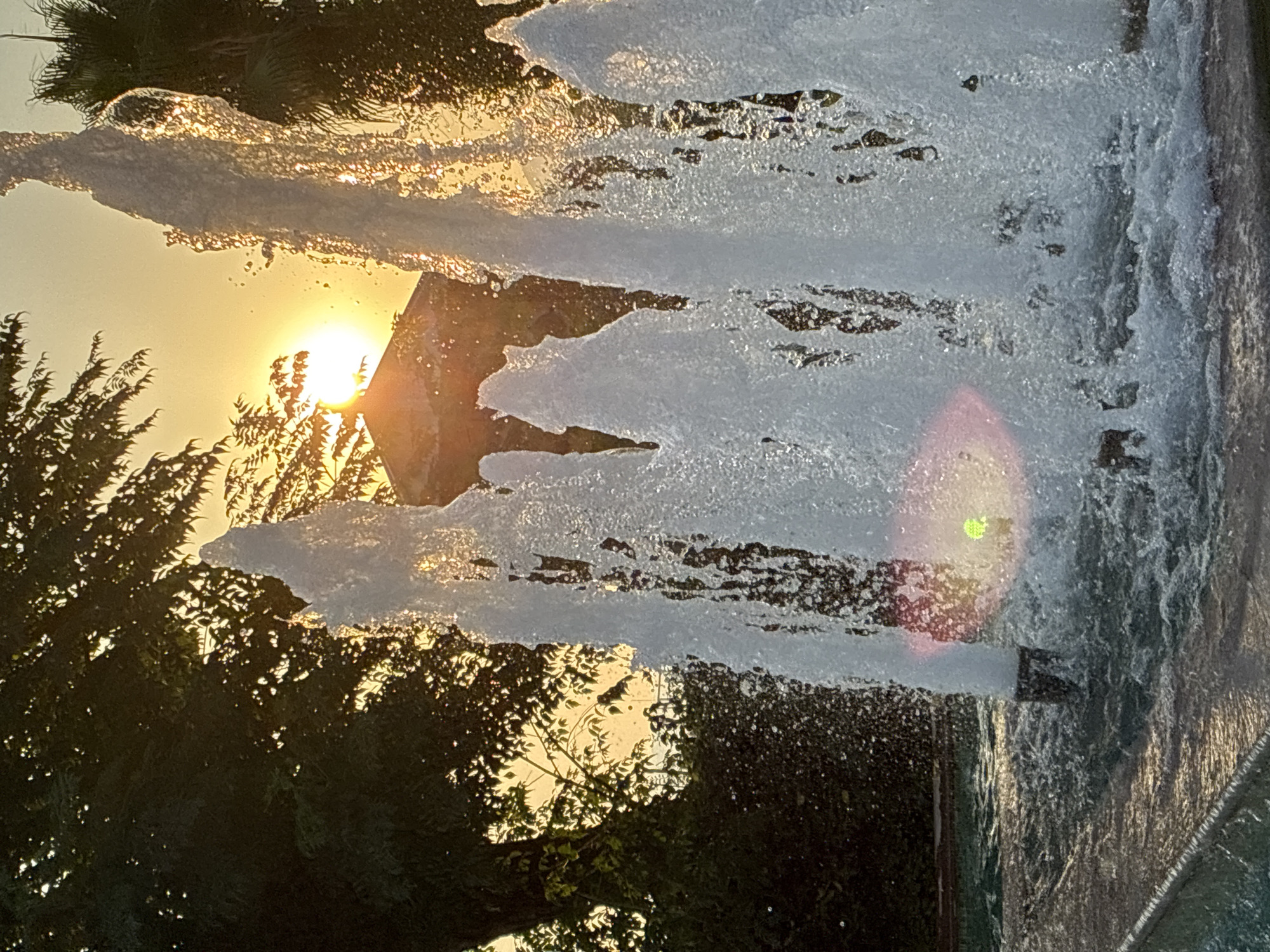 This media file is a photograph taken outdoors in Alanya, Turkey, during sunset. The image is vertically oriented and features a fountain with water spraying upwards. The sunlight is visible behind the water, creating a golden glow and highlighting the droplets. The sun is partially obscured by a tree with dark green foliage, and the leaves are silhouetted against the bright sky. In the background, a small part of a stone structure is visible, possibly part of the fountain or a nearby building. The water spray creates a curtain-like effect, with streaks of light and mist. A green lens flare is noticeable in the upper right portion of the image, indicating the direction of the light source. The overall impression is one of a serene and beautiful natural scene at dusk, with the warm light of the setting sun interacting with the water from the fountain.