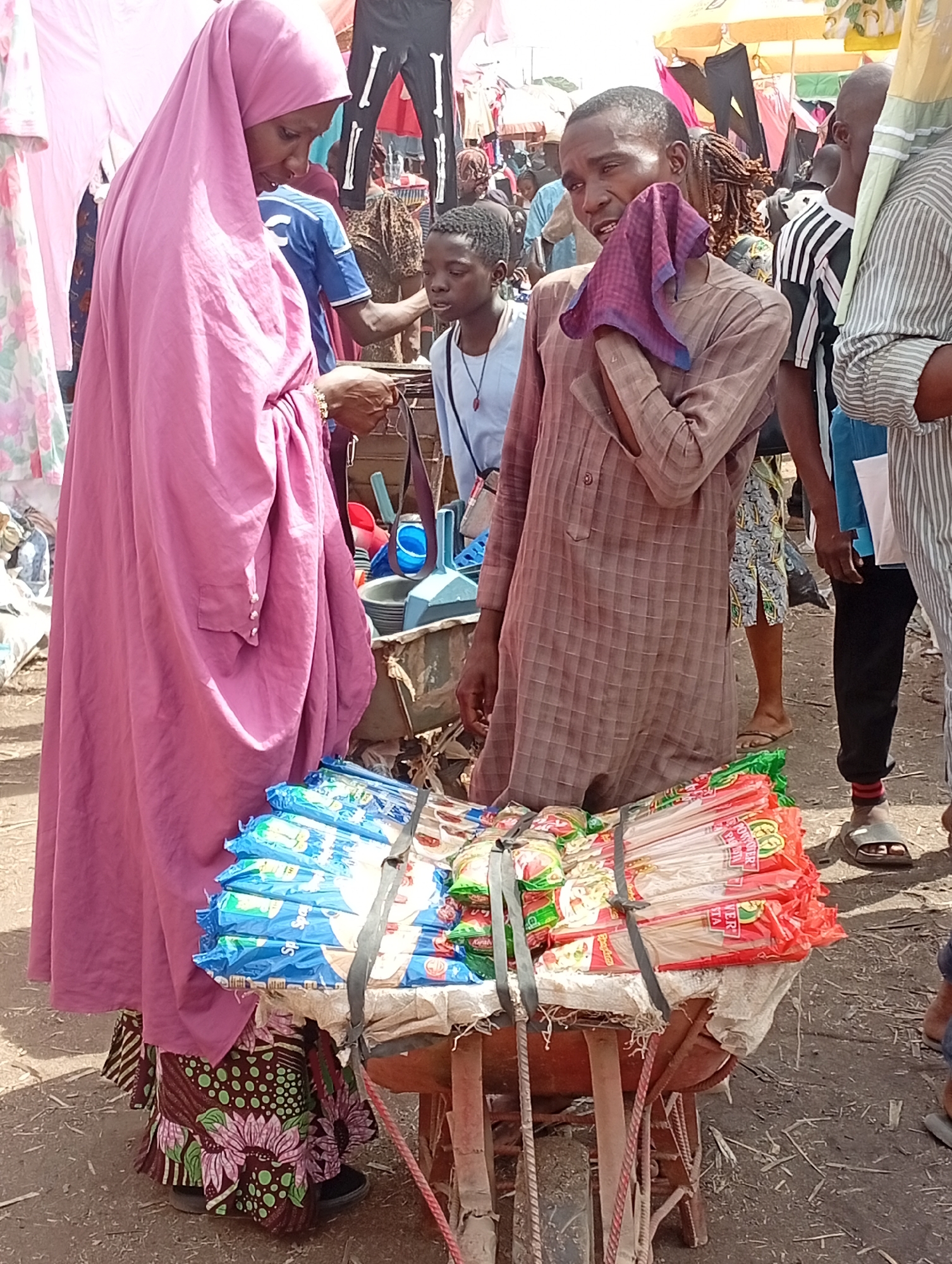 In an outdoor market scene in Kakuri Gwari, Nigeria, a woman clad in a floor-length pink hijab and a patterned skirt examines merchandise on a pushcart. She appears to be inspecting an item, potentially for purchase. The cart is loaded with neatly stacked packages of noodles, secured with black straps. A man, standing beside the cart, wipes his face with a purple cloth. He is wearing a brown checkered shirt. Behind them, other shoppers mill about, indicating a busy marketplace environment. Clothes hang in the background. The ground is dry and dusty.