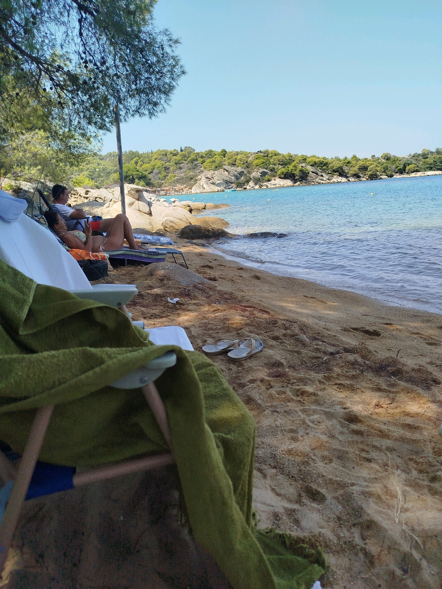 This is a horizontal, eye-level, medium shot of a beach in Sithonia Municipal Unit, Greece. The image shows a sandy beach with the sea on the right and people relaxing on chairs on the left. Two people are sitting on beach chairs, facing the sea. A man in a white t-shirt is on the left, and a woman in a bikini is on the right. A green towel is draped over a white beach chair in the foreground. The sea is a vibrant blue and the sky is clear and light blue. The background shows a rocky coastline covered in green trees. The overall scene depicts a relaxed day at the beach.