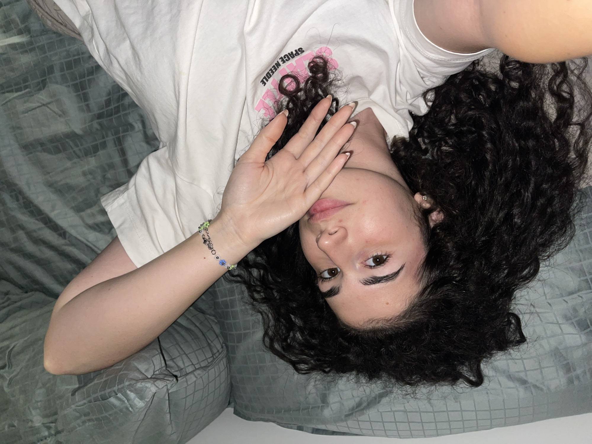 An overhead, slightly rotated shot captures a person, likely female, lying on their back on a textured dark teal or greyish-green blanket or bedspread with a grid pattern. The individual has dark, abundant curly hair spread around their head. They are wearing a white t-shirt that displays the pink text "SPACE NEEDLE" on the upper left chest area.
The person's left hand is raised, partially covering their mouth and nose, with fingers slightly splayed, revealing only their forehead, eyebrows, and one light brown eye looking directly at the camera. A colorful beaded bracelet with green, blue, and silver-toned beads is visible on their left wrist. Their fingernails have white tips. The lighting suggests an indoor environment. The person's visible eye conveys a neutral or slightly curious expression. Based on provided context, the image is situated in Sofia, Bulgaria.