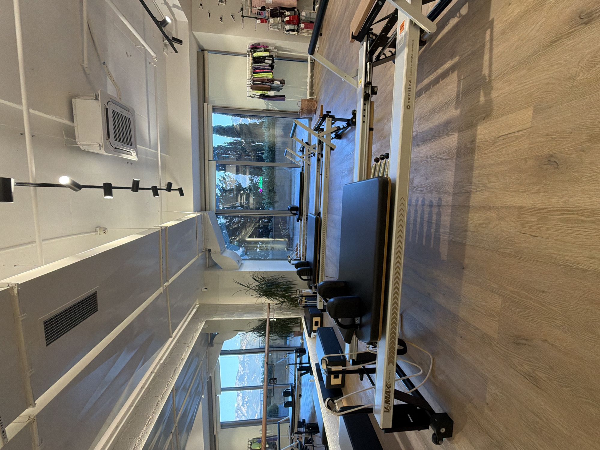The image depicts an indoor pilates studio, likely located in Boreti, Montenegro, with multiple reformer machines lined up. Large windows offer a view of trees and distant mountains, suggesting an outdoor environment visible from the studio. The studio features light-colored wooden flooring and a white ceiling with exposed beams and ventilation systems. Several pilates reformers, predominantly black and silver with some wooden accents, are the main focus. The machines are arranged in parallel rows. In the background, a rack of athletic wear is visible, along with some potted plants. The overall atmosphere is clean and modern, suggesting a professional fitness facility. There are no people visible in the image.