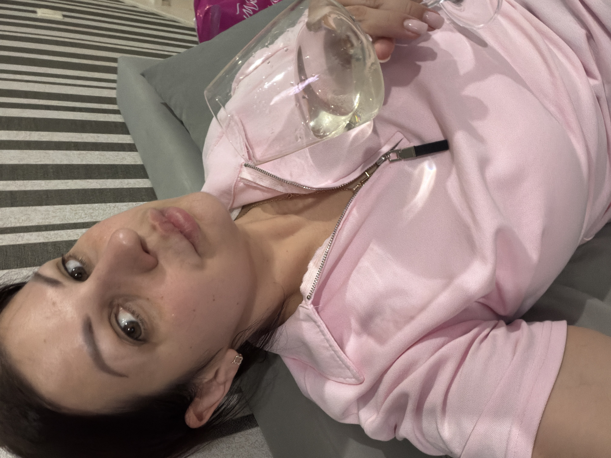 A woman with dark hair is pictured from the chest up, holding a glass of white wine. She is wearing a pink collared shirt with a zipper. The background consists of a gray couch and a wall with vertical black and white stripes. This image was taken in Sochi, Russia.