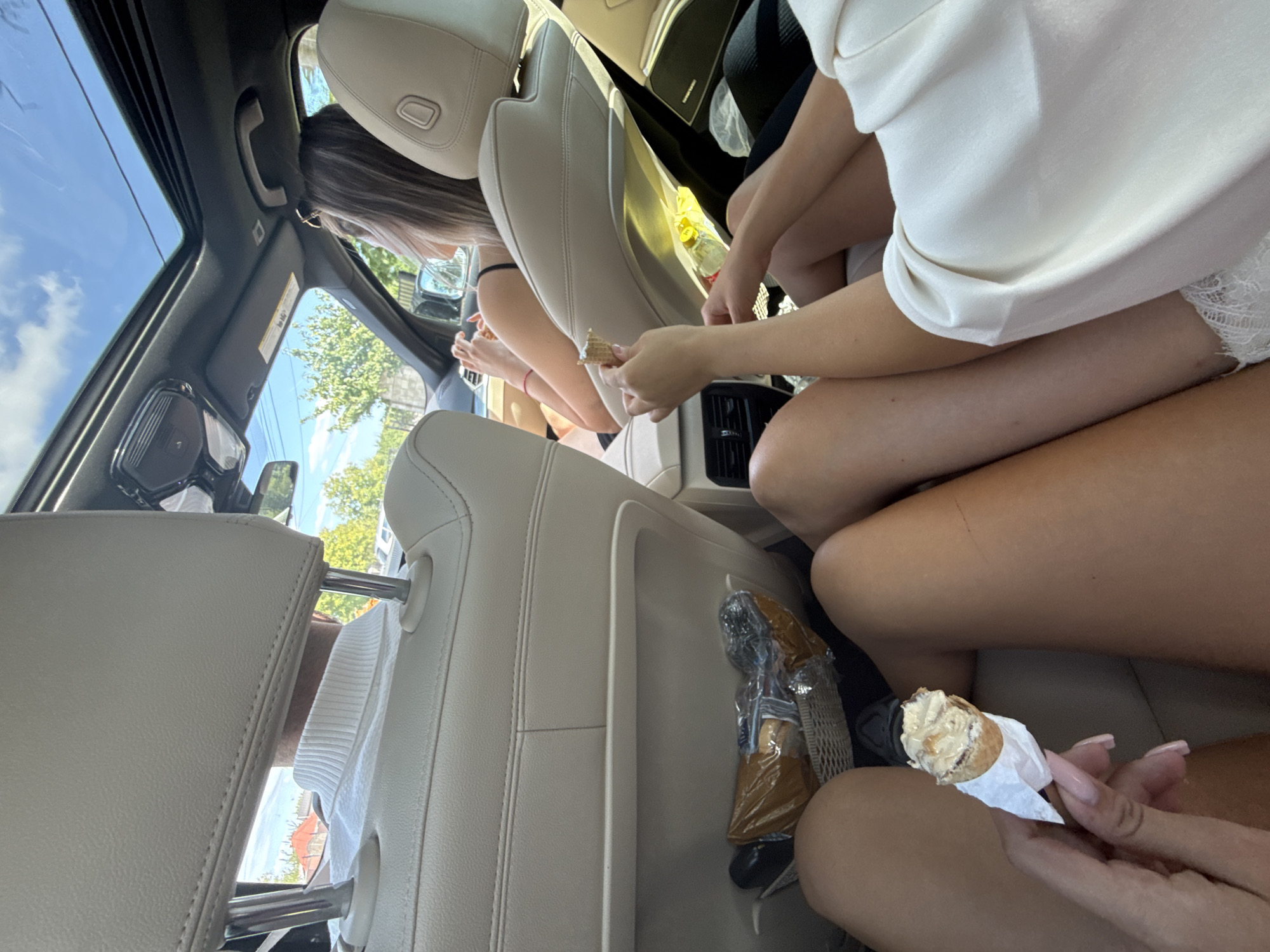 The image depicts the interior of a car with a sunroof open on a sunny day. In the foreground, the legs of at least three individuals are visible, seated in the back. One person is holding a partially eaten ice cream cone. In the middle ground, the back of a man's head wearing a white turtleneck is visible, and further back, a woman with blonde hair is seen driving. A bag containing food items is placed in a seat pocket. The car appears to be traveling on a road, with trees and buildings visible through the windows, suggesting an urban or suburban setting. There is no specific visual information within the image that definitively places the scene in Bogdănești, Romania.