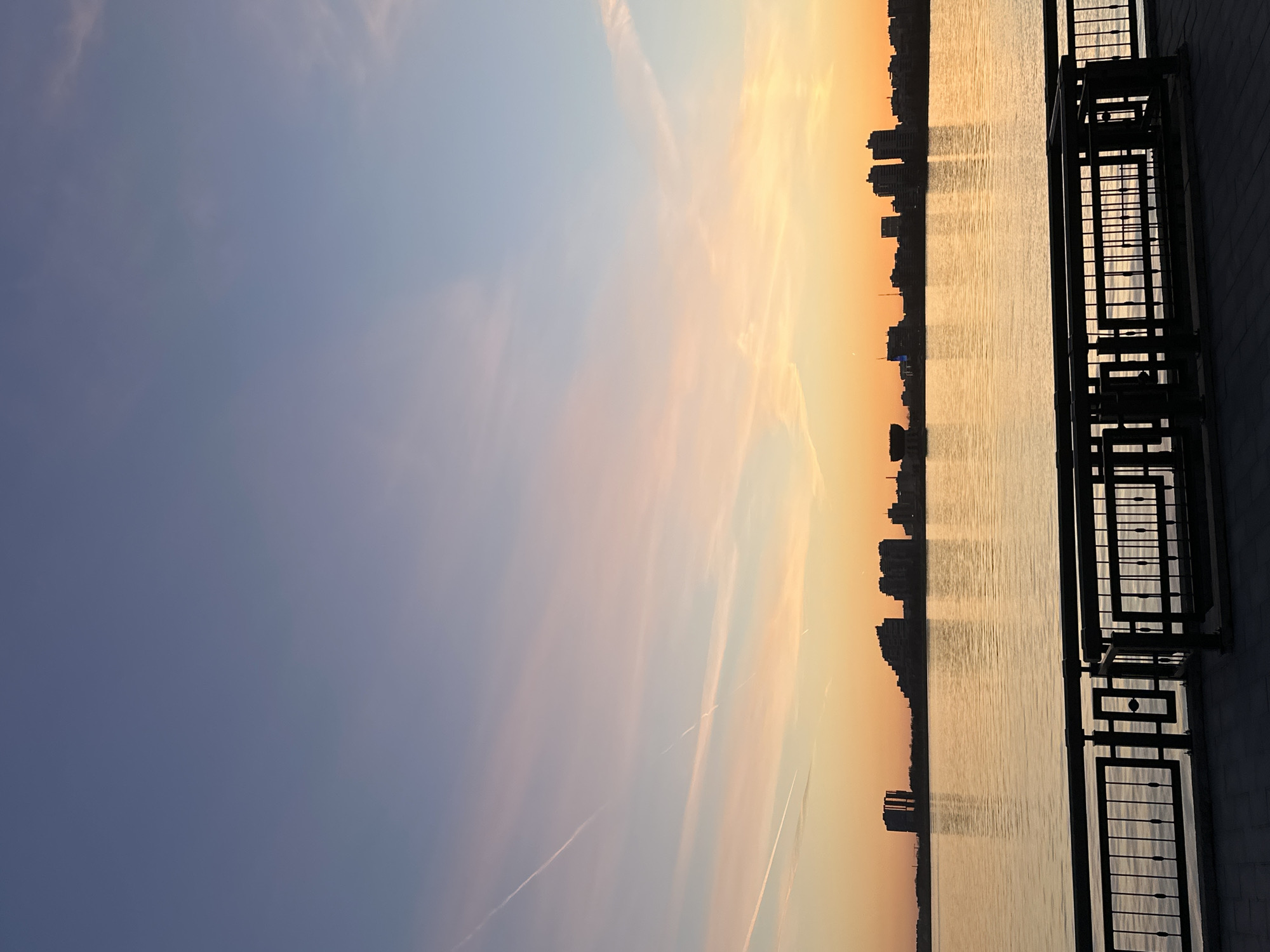 The image depicts a serene waterfront cityscape at sunset or sunrise. When viewed in a landscape orientation, the top half of the frame is dominated by a clear sky, exhibiting a beautiful color gradient from soft blue and light purple on the left to warm oranges and yellows on the right, indicating the sun's low position. Several thin, elongated clouds and faint airplane contrails are visible against the gradient.
A wide body of water occupies the middle ground, reflecting the sky's golden light and showing subtle ripples across its surface. Along the far bank of the water, a cityscape is presented in silhouette, with the dark outlines of numerous buildings stretching across the horizon, characteristic of an urban environment.
In the immediate foreground on the right side, a dark, decorative metal railing with geometric designs runs vertically. Behind it, a dark, solid wall is partially visible, suggesting the perspective is from a promenade or viewing platform overlooking the water. No specific activities or people are present, and there is no visible text. The overall impression is one of peaceful twilight over Kazan, Russia.
