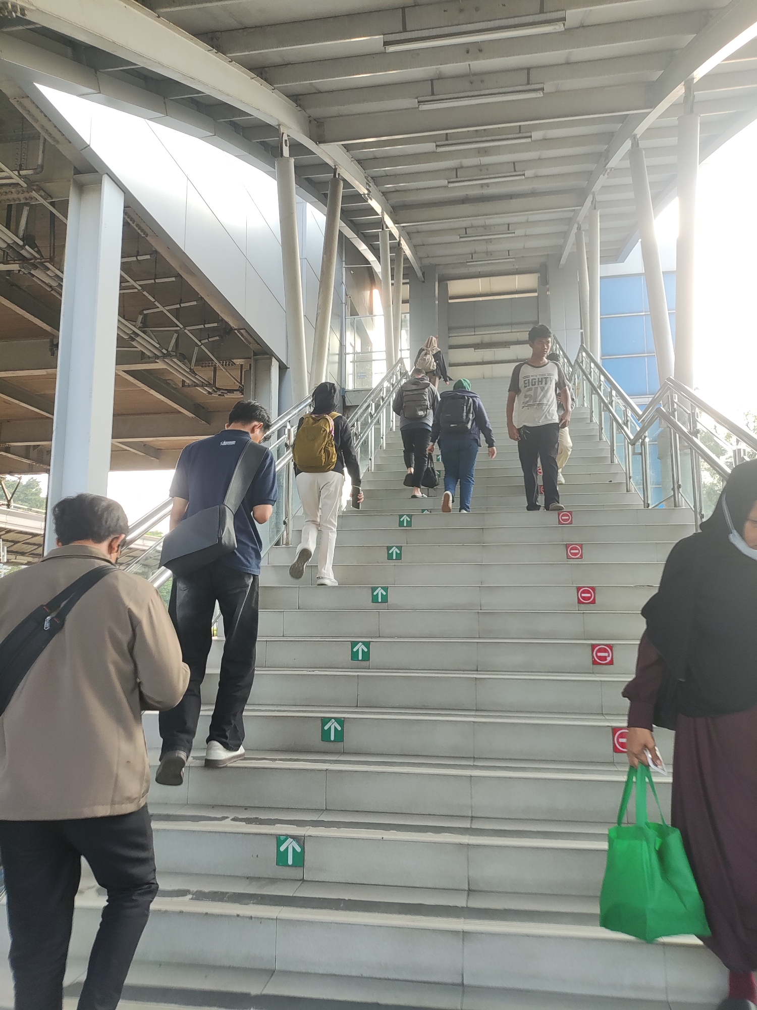 The image depicts an outdoor stairway at a transportation hub in East Jakarta, Indonesia, likely a train station or elevated walkway. Several individuals are ascending the wide, concrete stairs. A person in a brown jacket with a black bag slung over their shoulder is walking up, slightly ahead of a man in a dark blue polo shirt and black pants, also with a bag. Further up the stairs, a woman with a backpack and light-colored pants is walking, followed by another person with a backpack. At the top of the visible stairs, a young man in a white t-shirt with "EIGHT" printed on it and black pants is walking upwards, alongside another person. A woman in a dark maroon dress and hijab, carrying a bright green reusable bag, is also visible on the stairs, walking upwards. The stairway has metal railings on both sides and a metal overhead structure. Red circular signs with a horizontal line indicating "no entry" or "forbidden" are affixed to some of the steps, while green square signs with white upward arrows indicate the direction of ascent. The lighting suggests it is daytime, possibly late afternoon, with bright sunlight creating some glare. The overall environment is clean and modern, characteristic of urban infrastructure in Indonesia.