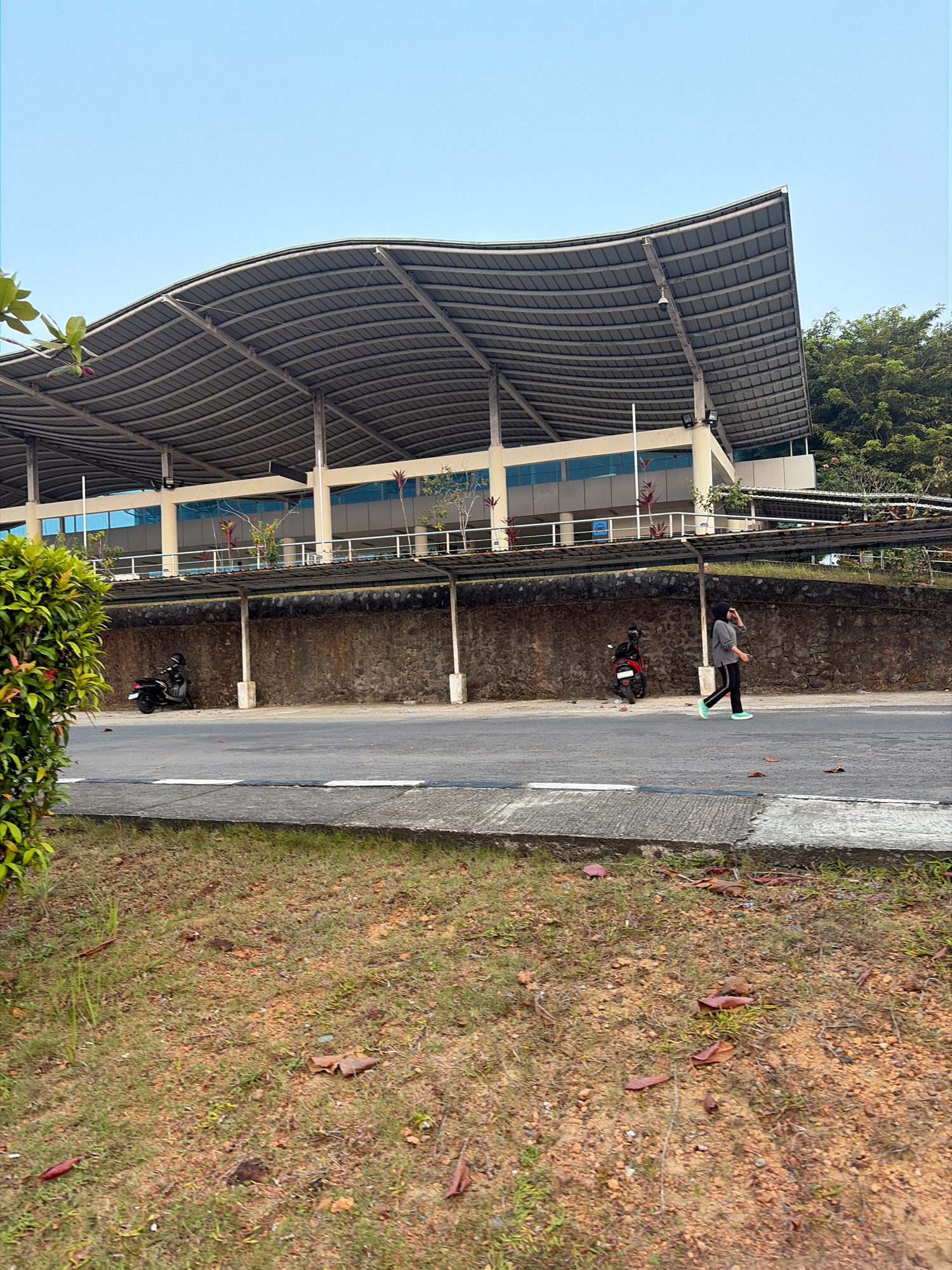 In Tanjung Pinang, Indonesia, a person wearing a grey hijab and athletic attire walks on a paved road in front of a building. The building features a distinct, wave-like metal roof supported by white pillars. Below the main structure, a concrete wall runs horizontally, with two motorcycles parked against it. The foreground consists of a grassy embankment with scattered dry leaves. The sky is clear blue, and trees are visible in the background, to the right of the building.