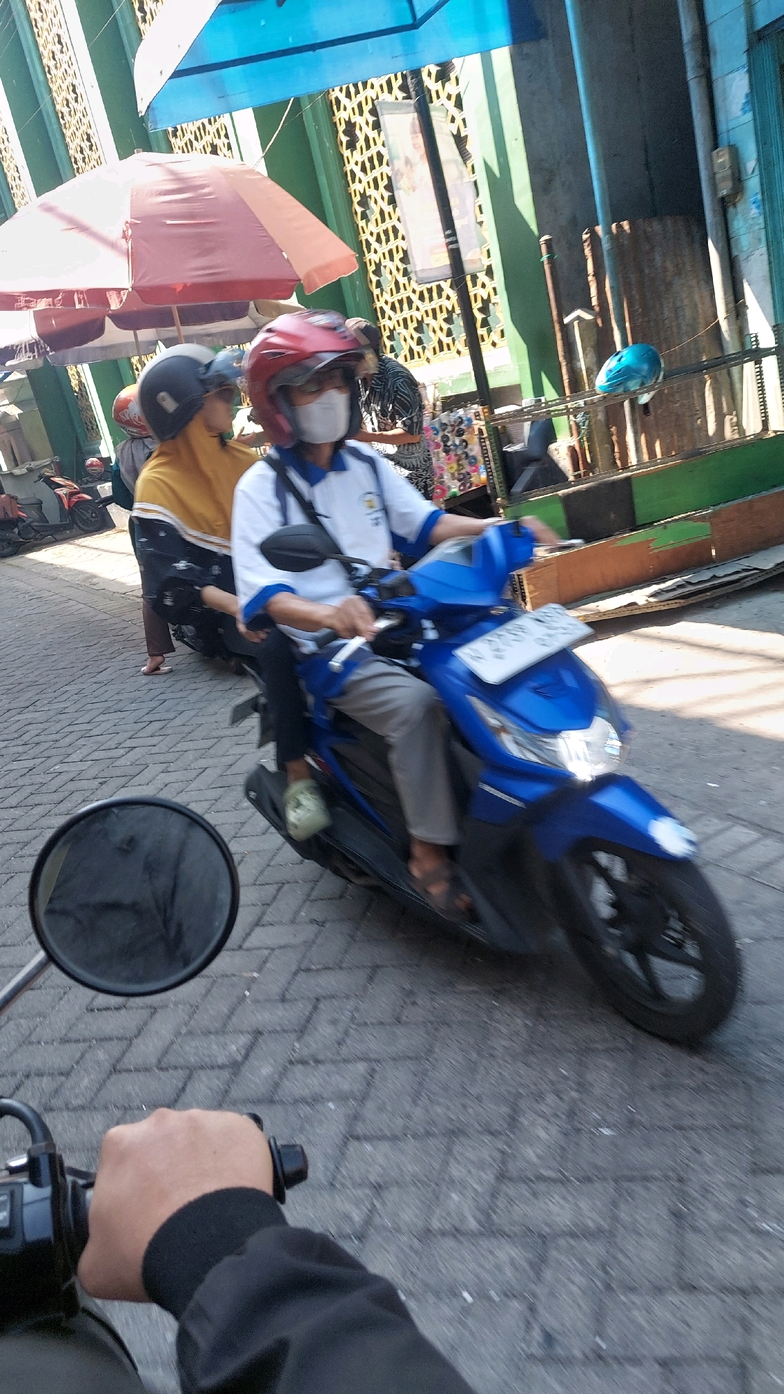 In a street scene in Sidoarjo, Indonesia, two individuals are riding a blue motorcycle. The person in front, wearing a red helmet and a white shirt with blue trim, is driving. They are also wearing a white face mask and grey pants. The person behind them, partially visible, is wearing a yellow hijab and a black and white patterned garment. The motorcycle is on a brick-paved street, with a red umbrella and a blue tarp visible in the background, suggesting a market or roadside stall area. Sunlight casts shadows on the street, indicating it is daytime. Other motorcycles and a blue helmet are also present in the background.
