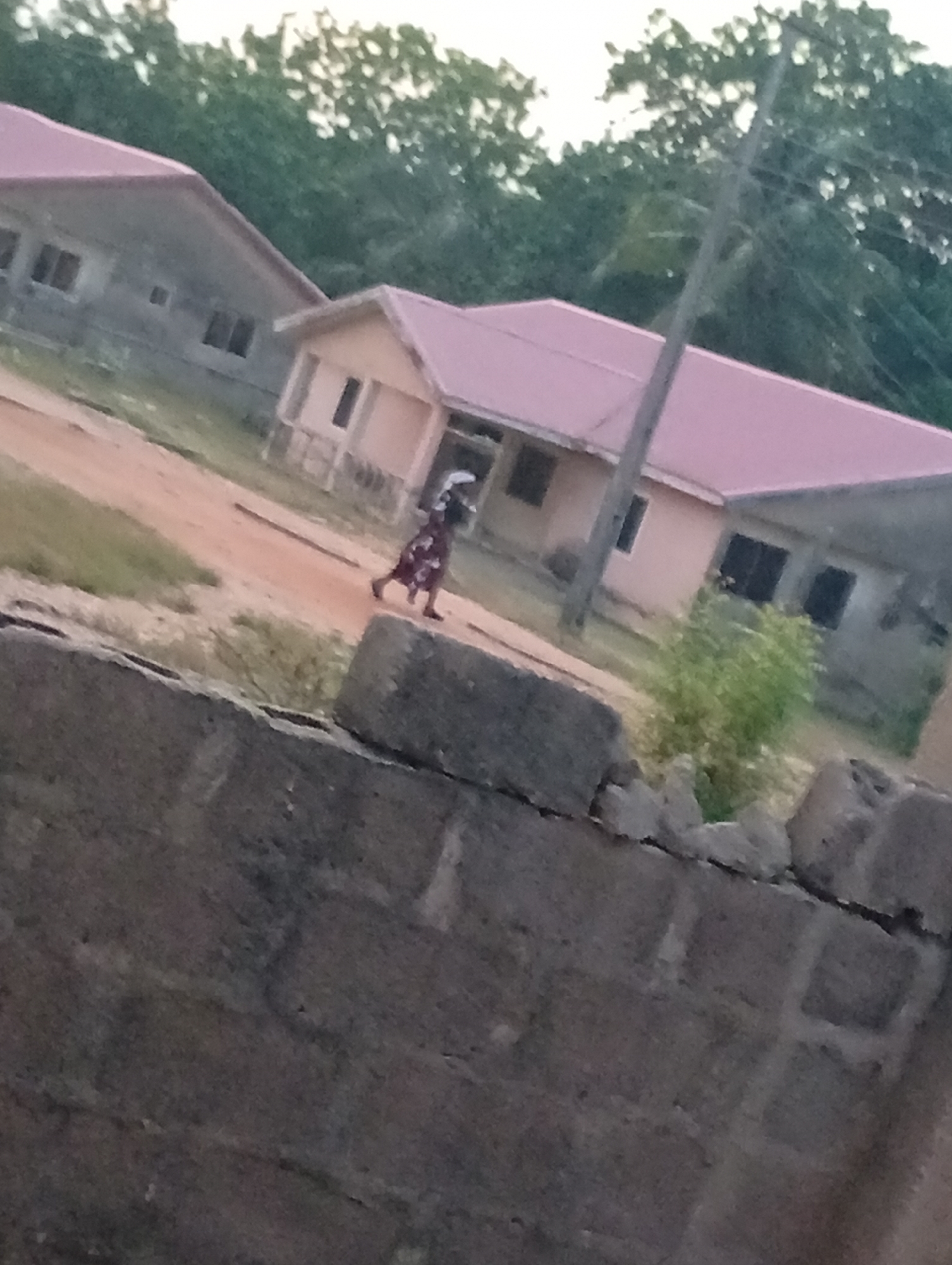 A low-angle shot captures a scene with a partial view of a concrete block wall in the foreground. Behind the wall, a dirt road leads to a residential area with several single-story buildings that have pink roofs. A person is walking on the road. A utility pole is visible in the background and green trees are in the very back. The location appears to be in an area with modest housing and minimal infrastructure.
