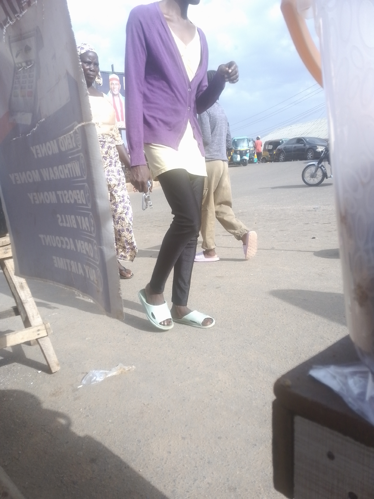 The media file shows an outdoor scene with several people and vehicles. A person wearing a purple cardigan and dark leggings walks across the pavement. To their left, a woman in a patterned dress and headscarf is visible, looking towards the camera. In the background, several vehicles, including a blue and yellow auto-rickshaw, a black car, and a motorcycle, are on a road. The sky is cloudy, suggesting daytime. A large sign with reversed text in white lettering on a dark background is partially visible on the left side of the frame. The text on the sign appears to include phrases like "SEND MONEY," "DEPOSIT MONEY," "PAY BILLS," and "OPEN ACCOUNT," indicating it might be an advertisement for a financial service. The overall setting appears to be a street or market area, possibly in a town or city, given the presence of vehicles and commercial signage. The lighting suggests it is daytime, and the cloudy sky indicates moderate weather.