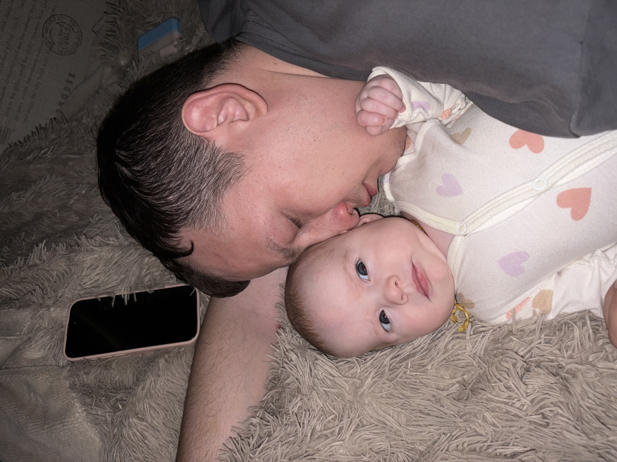 This media file features a man and a baby lying intimately together on a soft, shaggy grey blanket or rug, likely indoors in a home setting in Chaikovsky, Russia. The man, positioned on the left, has short, dark hair and light stubble, wearing a dark grey t-shirt. He is leaning in towards the baby, with his nose gently touching the baby's, suggesting a tender kiss or affectionate nuzzle.
The baby, on the right, has fair, fine hair and bright blue eyes, looking directly at the viewer with a curious and slightly smiling expression. The baby is dressed in a light-colored onesie decorated with scattered pink, purple, and orange heart patterns. A small, yellowish beaded necklace, possibly an amber teething necklace, is visible around the baby's neck. The baby's tiny fist is tucked near the man's arm.
In the lower left of the frame, a smartphone with a pink or rose gold case is lying screen-down on the blanket next to the man's arm. In the upper left background, behind the man's head, a small blue object, possibly a stapler or a box, is visible. Faint, dark text is printed on the blanket/rug in this area, with fragments like "HOUSE", "STUDIO", and "2021" discernable, though most of the text is illegible.
The lighting is soft and warm, contributing to the cozy and loving atmosphere of the scene. The image conveys a strong sense of affection and connection between the man and the baby.
